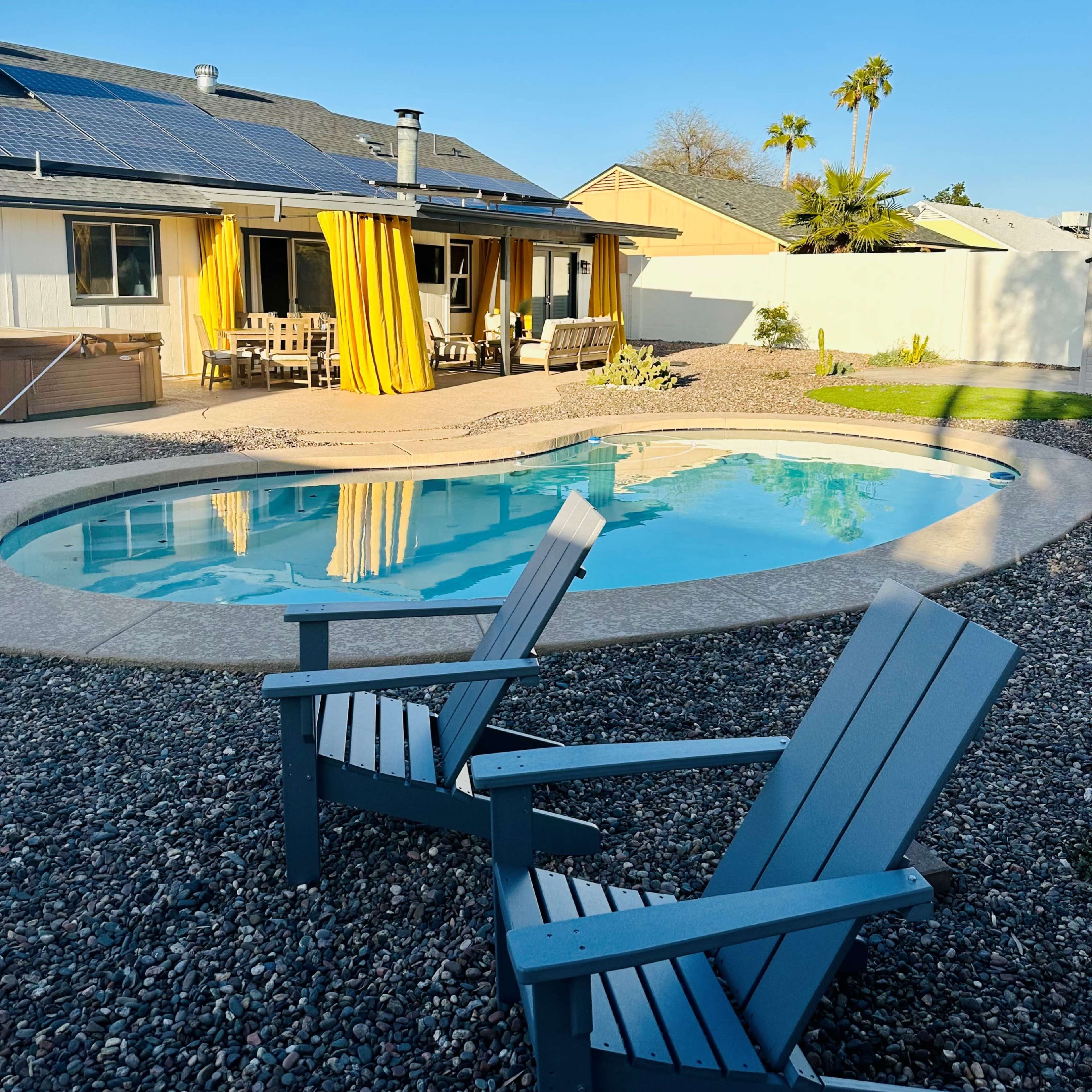 A blue swimming pool is surrounded by gravel and two gray adirondack chairs, with a house featuring solar panels and yellow drapes in the background.