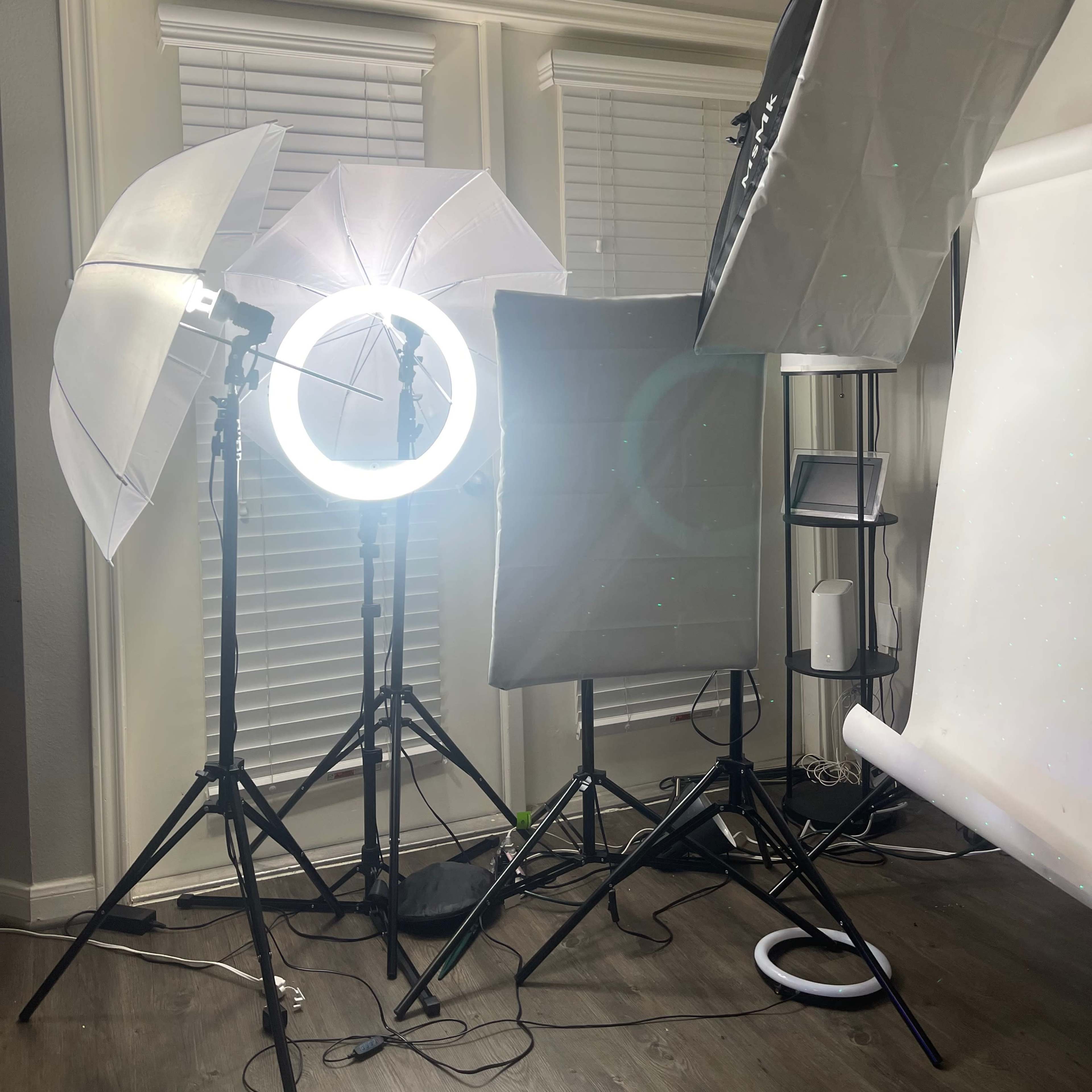 A photography setup featuring multiple light sources, including softboxes and a ring light, arranged around a backdrop in a room with white blinds.