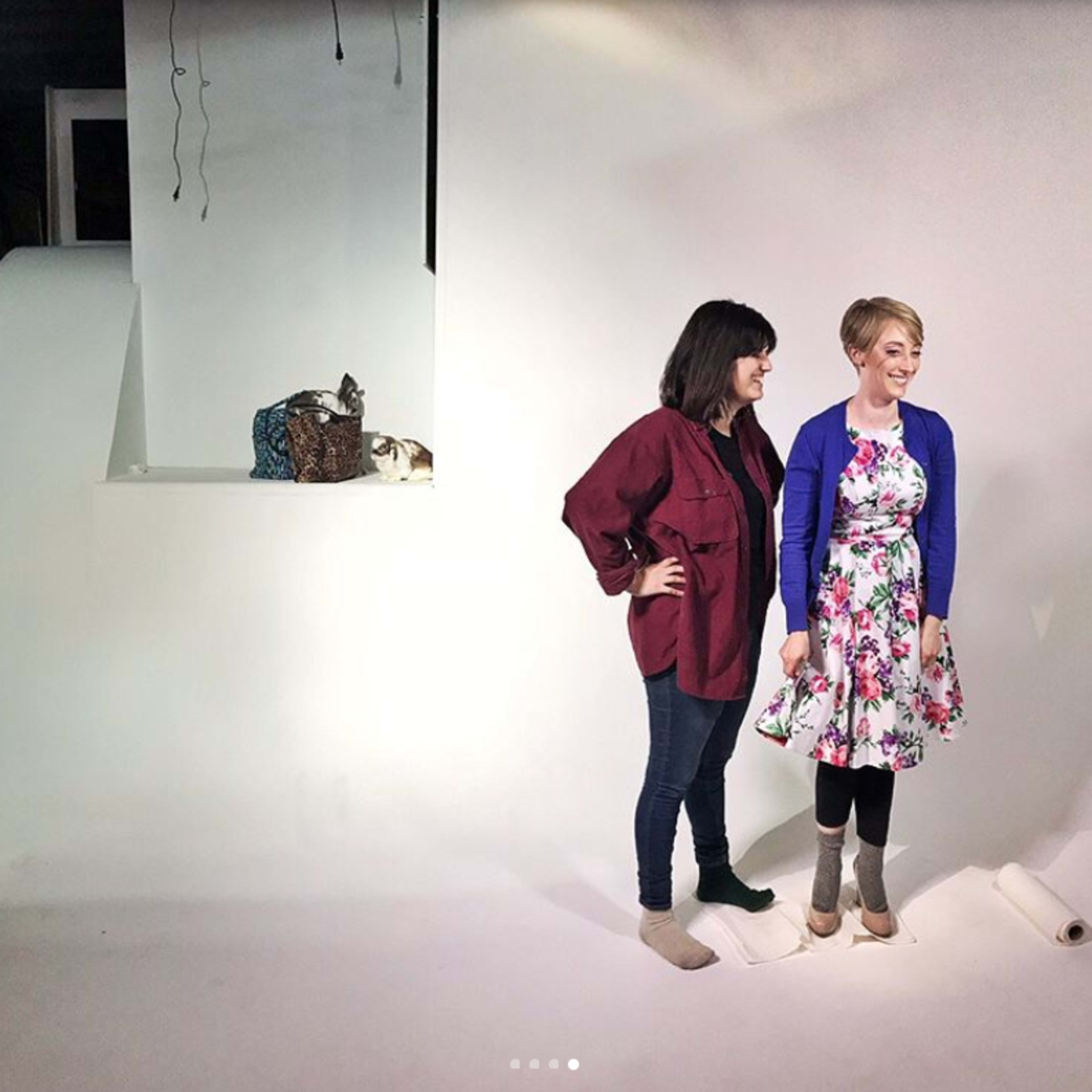 Two women stand on a white backdrop, one wearing a floral dress and the other in a maroon shirt, with a basket and a cat visible in the background.