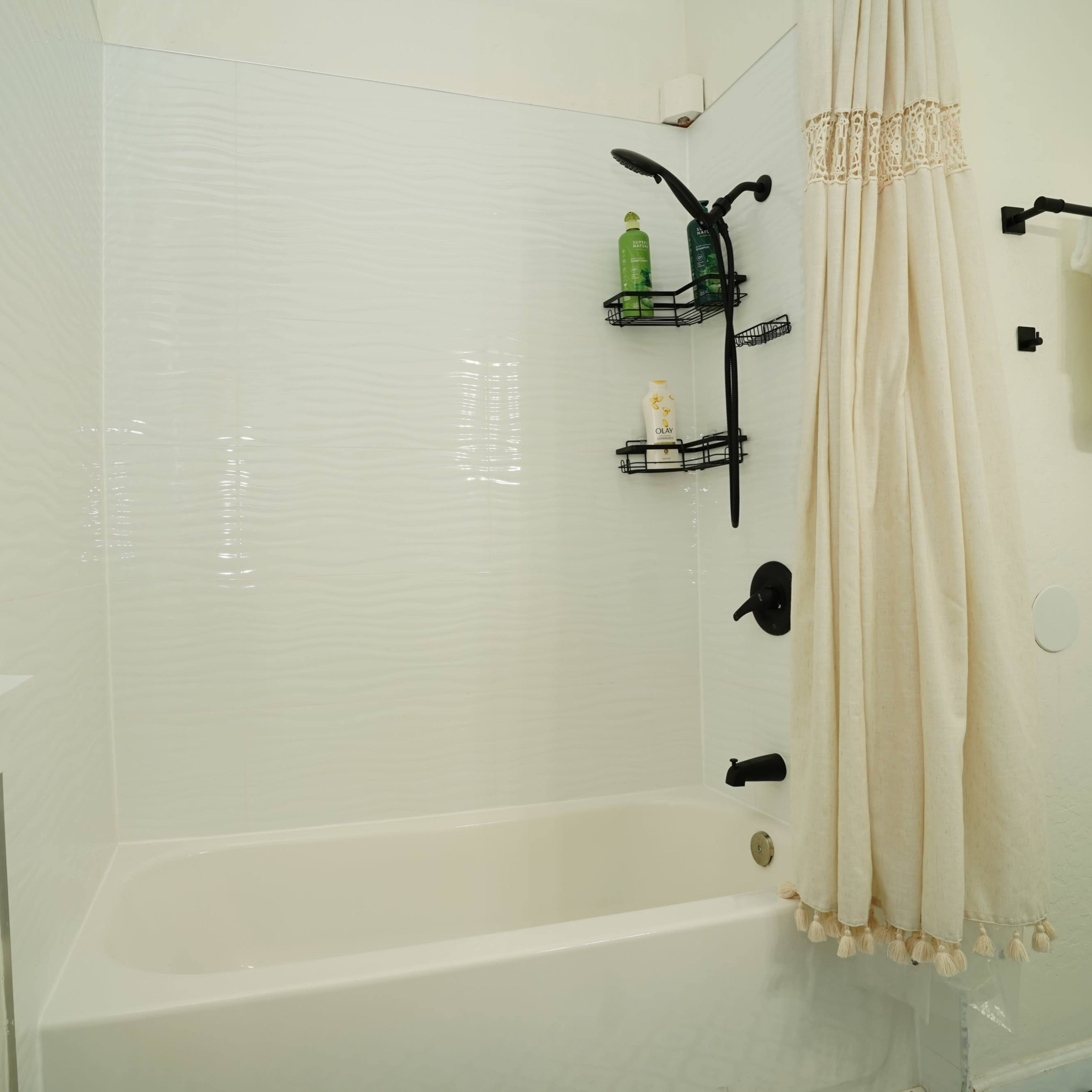 A clean, modern bathroom featuring a bathtub with a shower curtain, a wall-mounted showerhead, and a black shelf with toiletries.