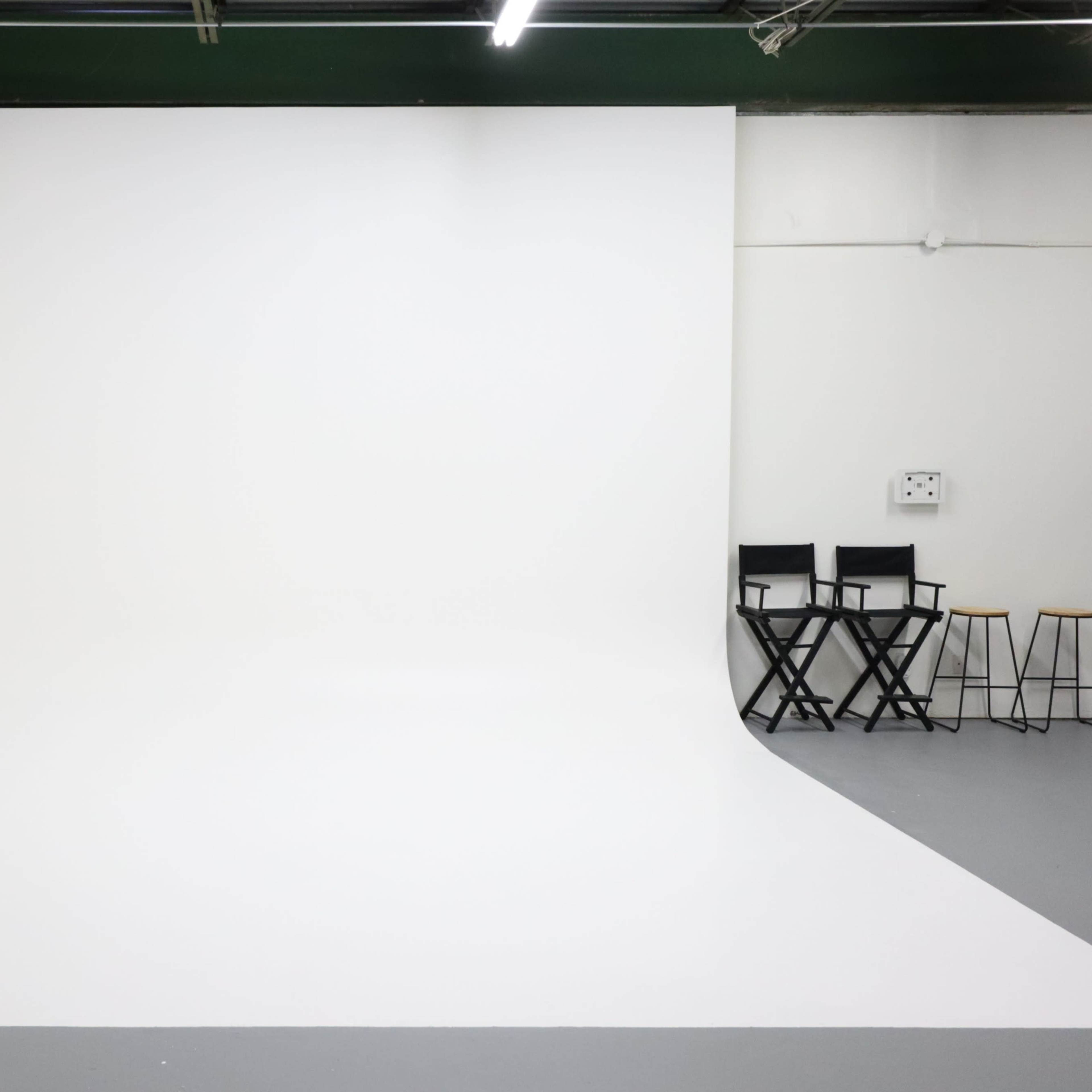 A minimalistic photography studio with a white backdrop and a few black chairs against a plain wall.