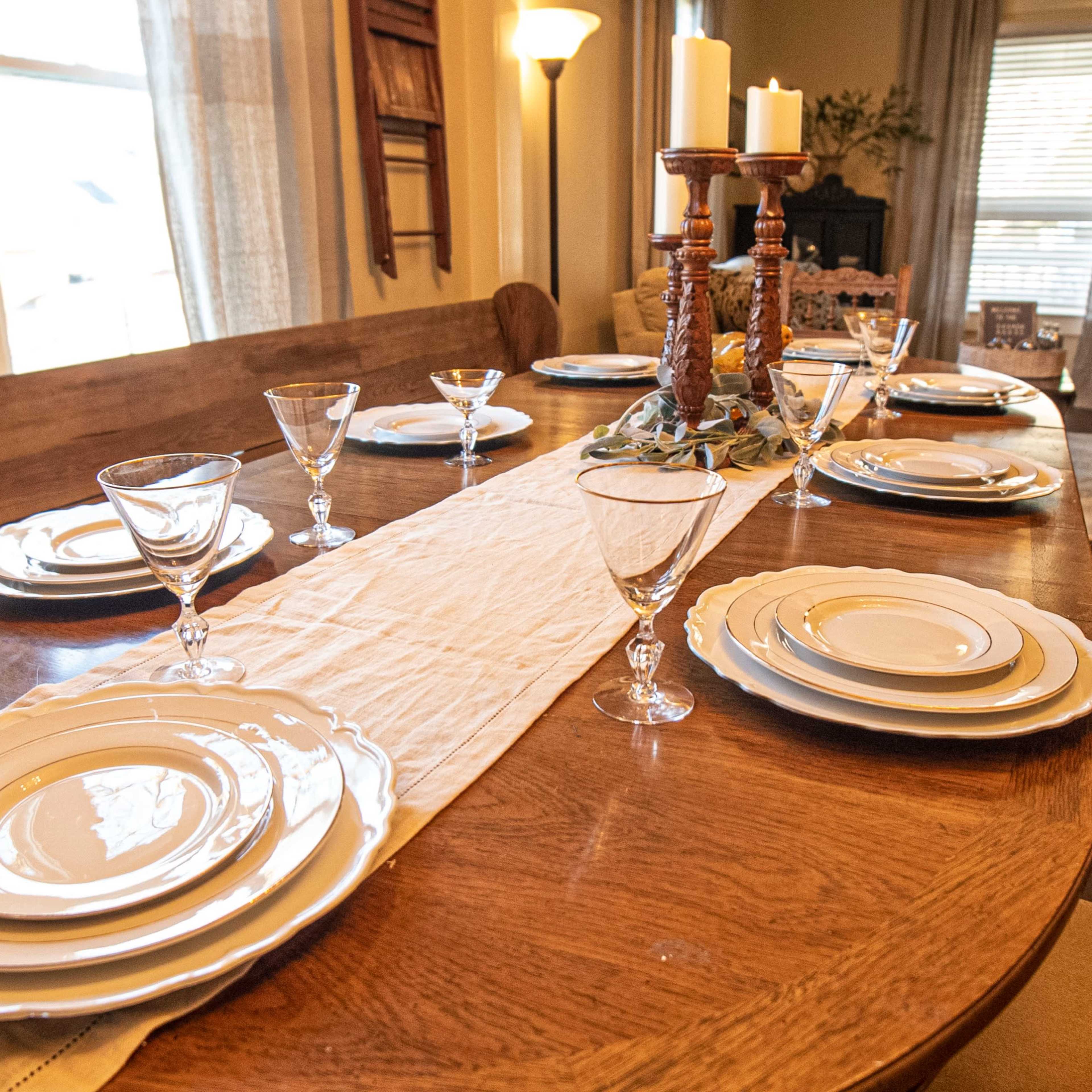 A wooden dining table is set with white plates, glassware, and a decorative runner, surrounded by chairs in a well-lit room.