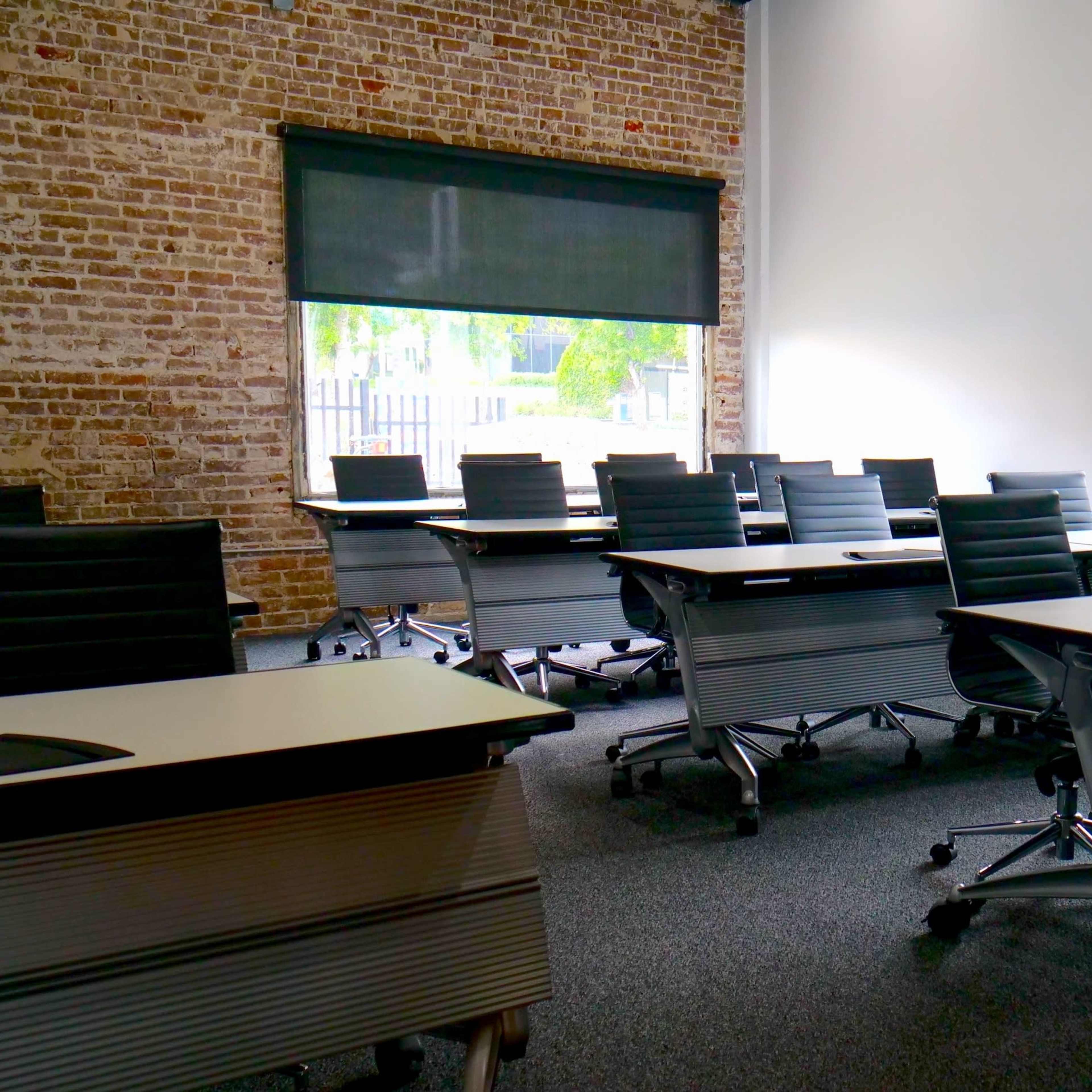 A modern conference room features several rows of tables and chairs, with large windows allowing natural light to enter through shades.