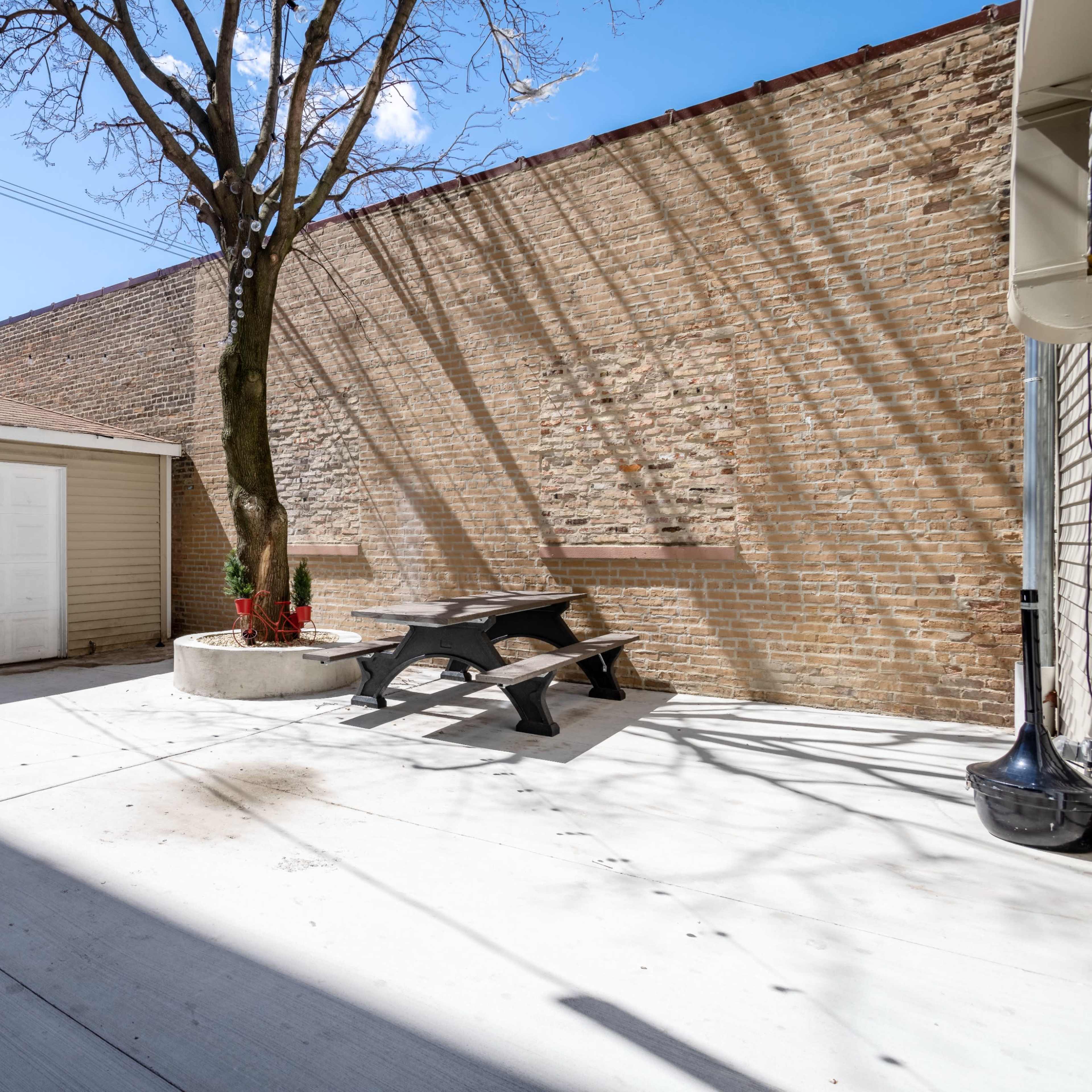 A paved outdoor area with a garage, a picnic table, and a tree in front of a brick wall under a clear sky.
