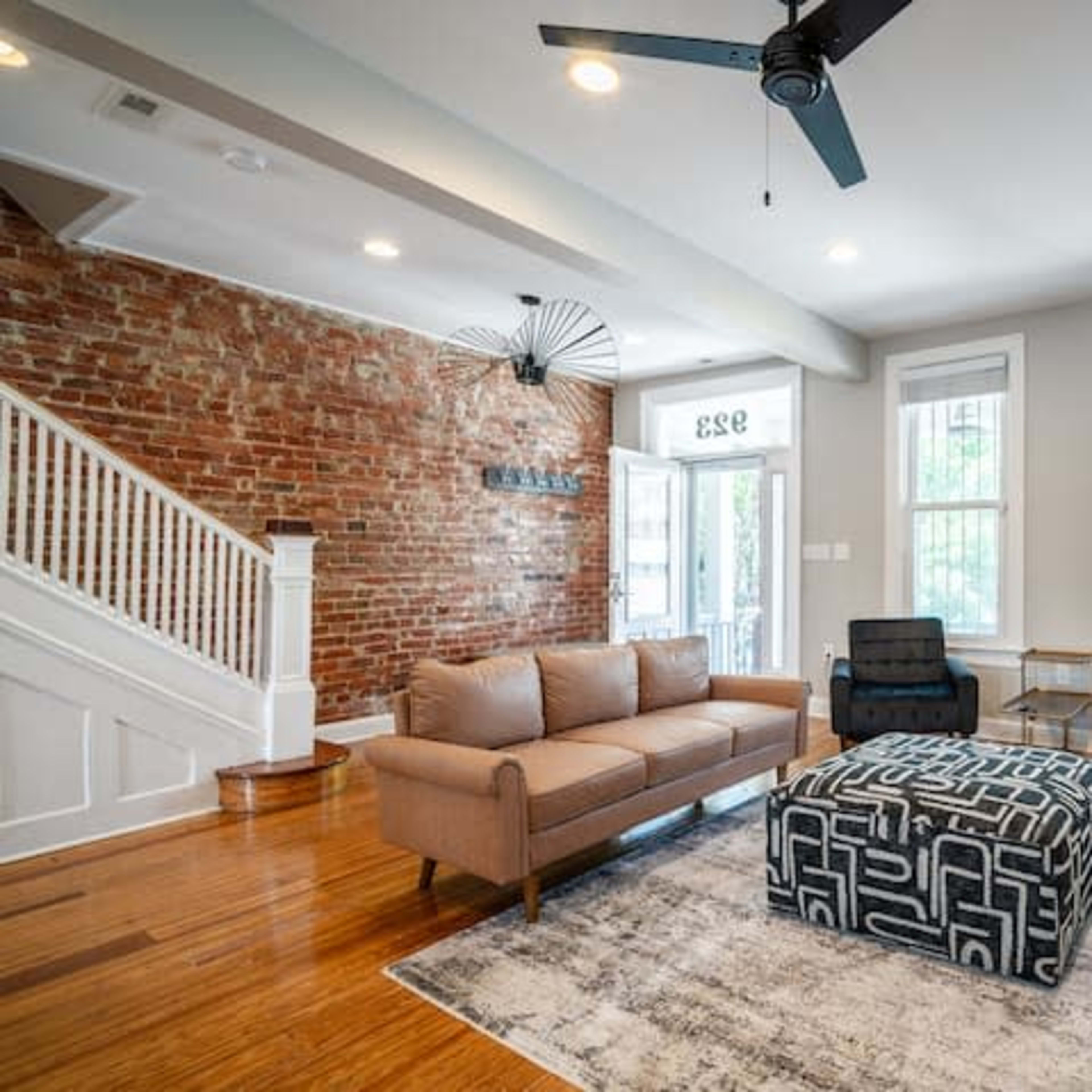 A living room featuring brick walls, a light-colored sofa, two black chairs, and a patterned ottoman on a wood floor.