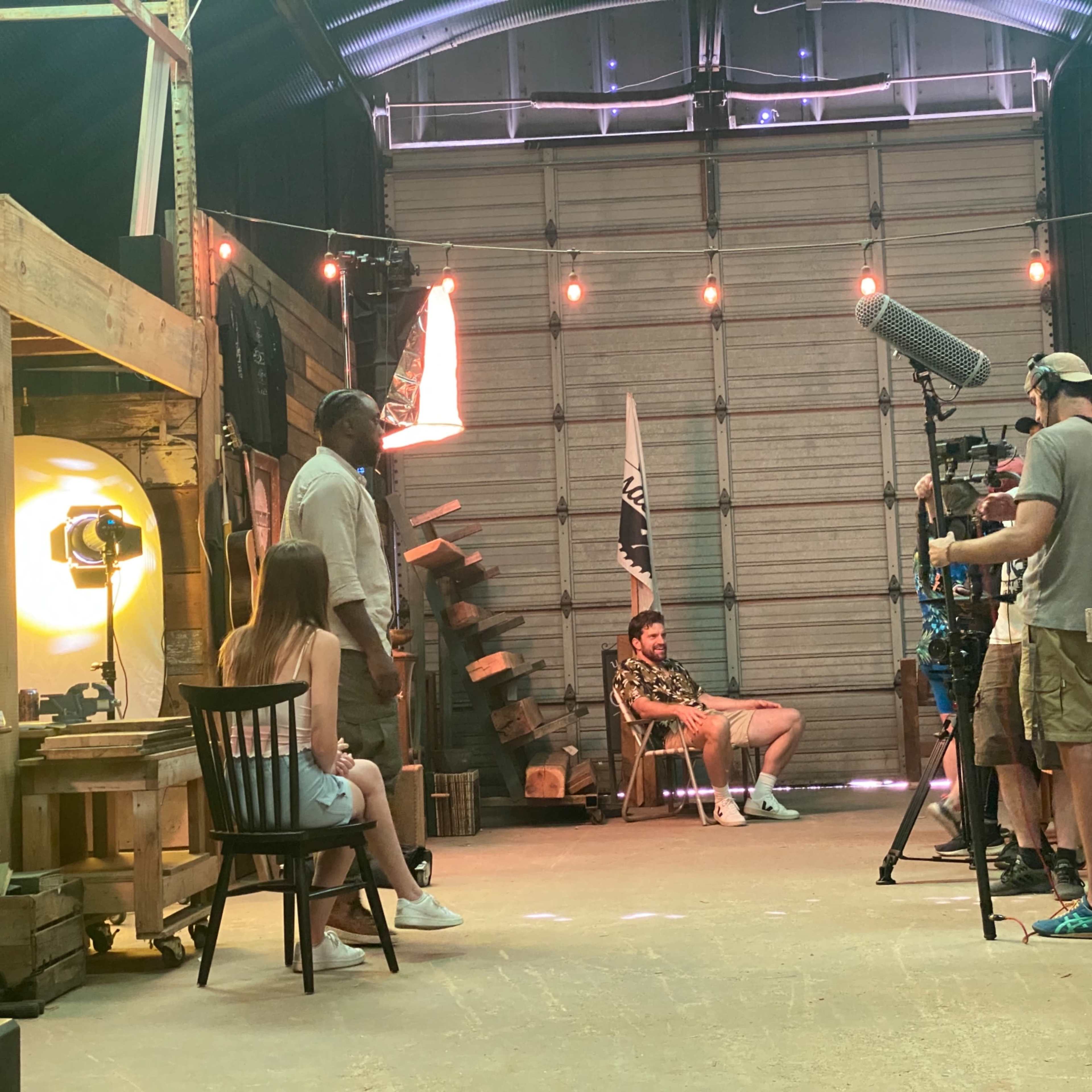A film crew sets up equipment in a workshop while two people are positioned for an interview.