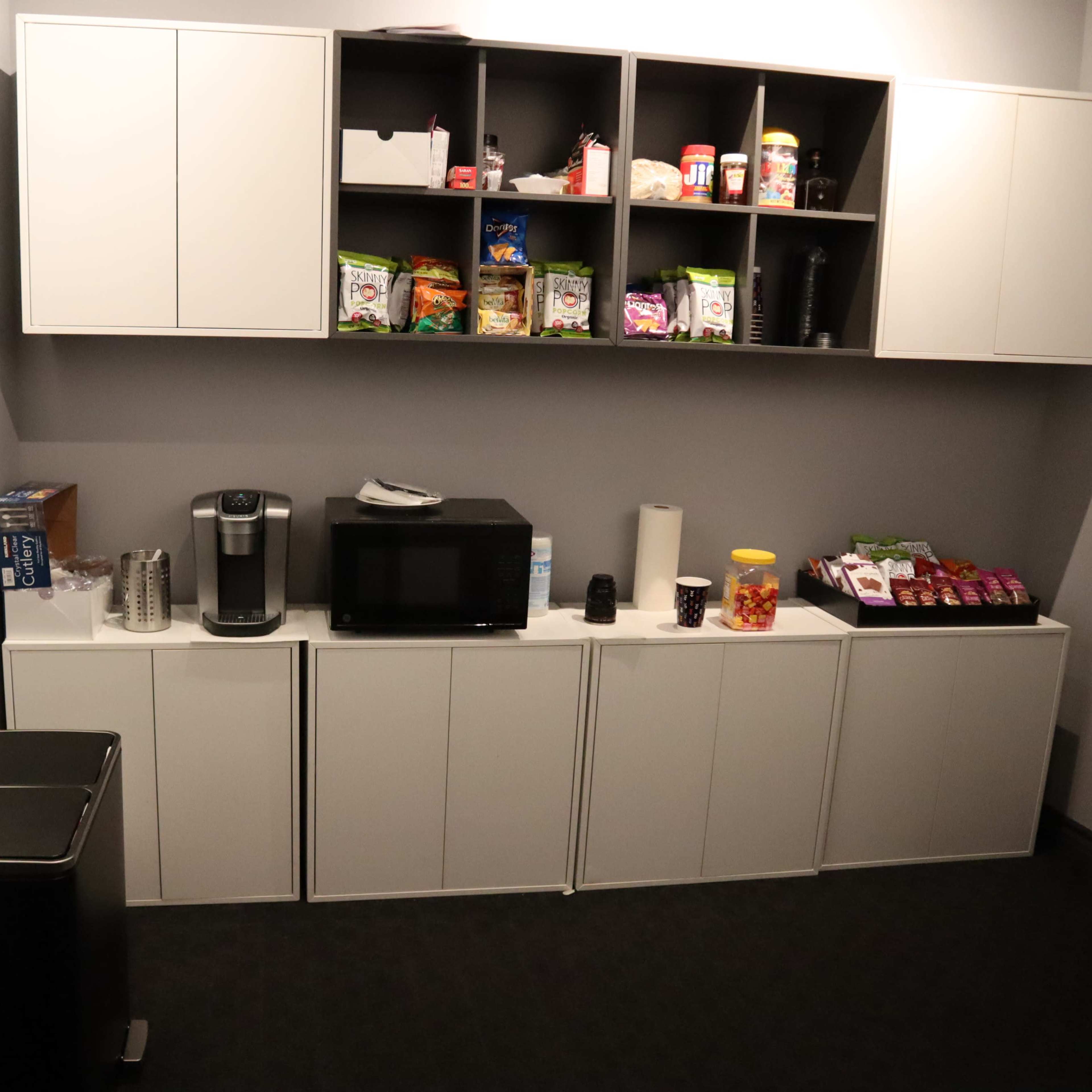 A small break room equipped with a microwave, coffee maker, a trash can, and shelves filled with snacks and drinks.