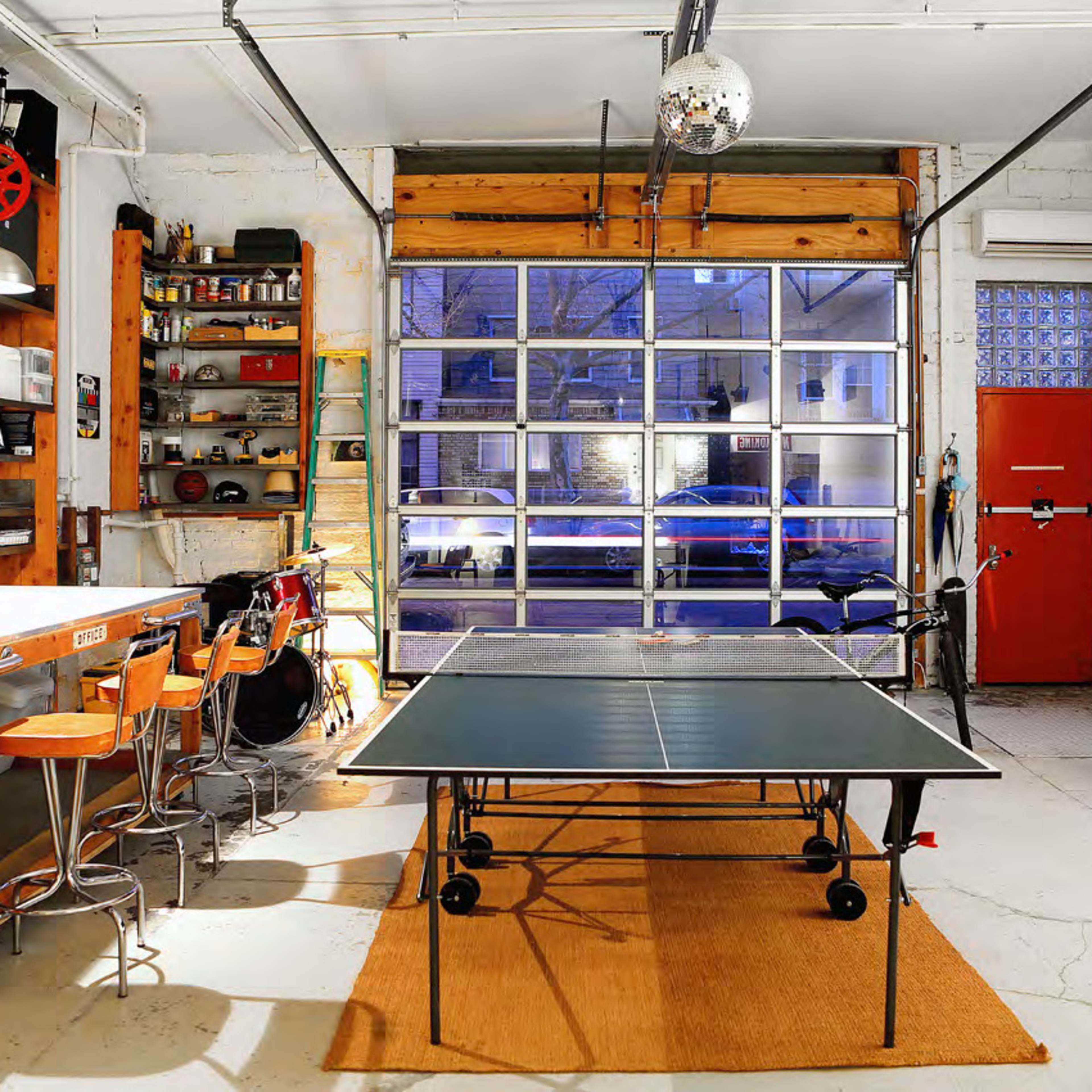 A modern garage space featuring a ping pong table, bar stools, shelving with tools and supplies, and a large garage door open to a view of an outdoor area.