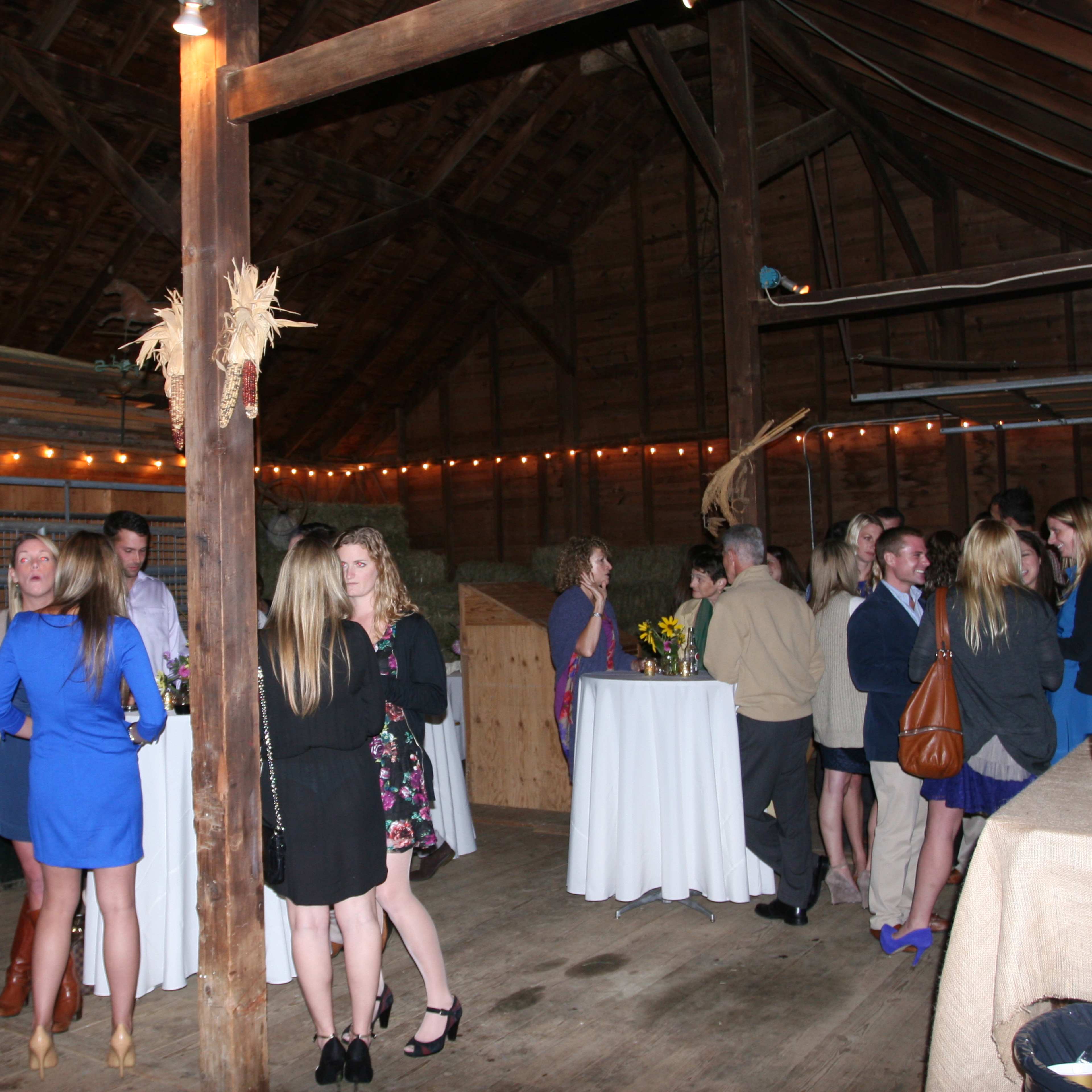 A group of people mingles at a social event inside a rustic barn, surrounded by tables and decorative lighting.