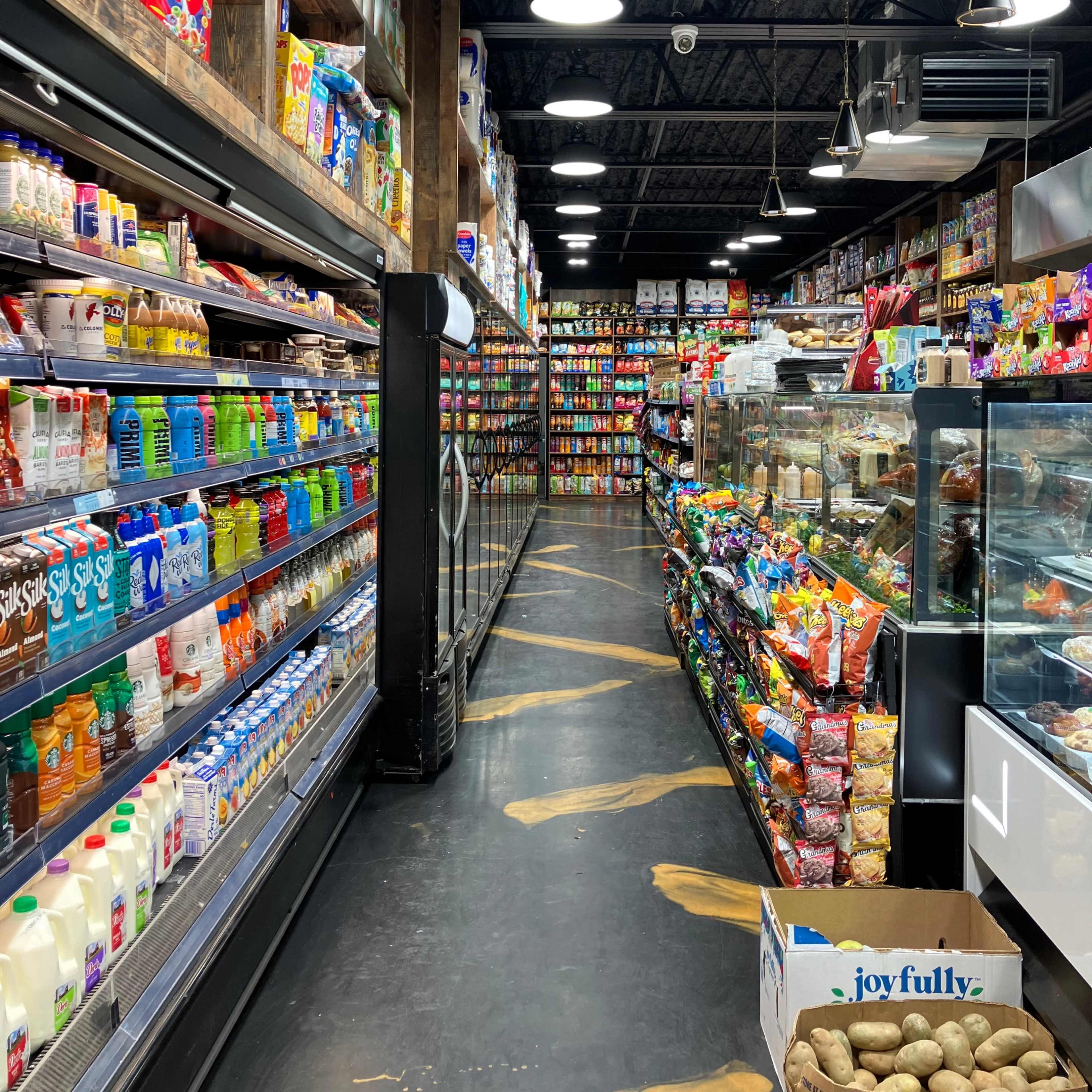 An aisle in a grocery store lined with shelves of colorful packaged goods, dairy products, and snacks, with a display case of food on one side.