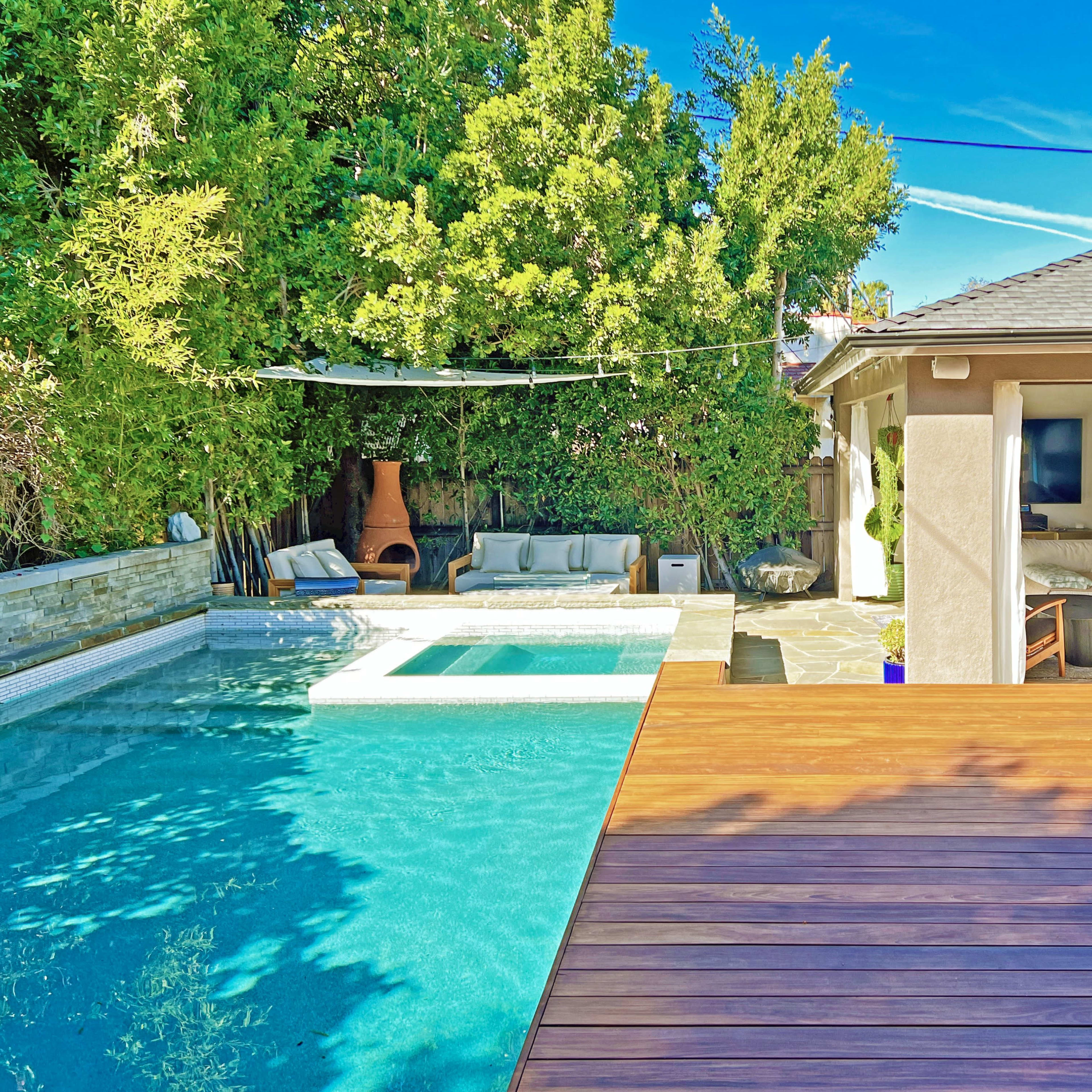 A modern backyard with a swimming pool, a wooden deck, and a covered seating area surrounded by greenery.