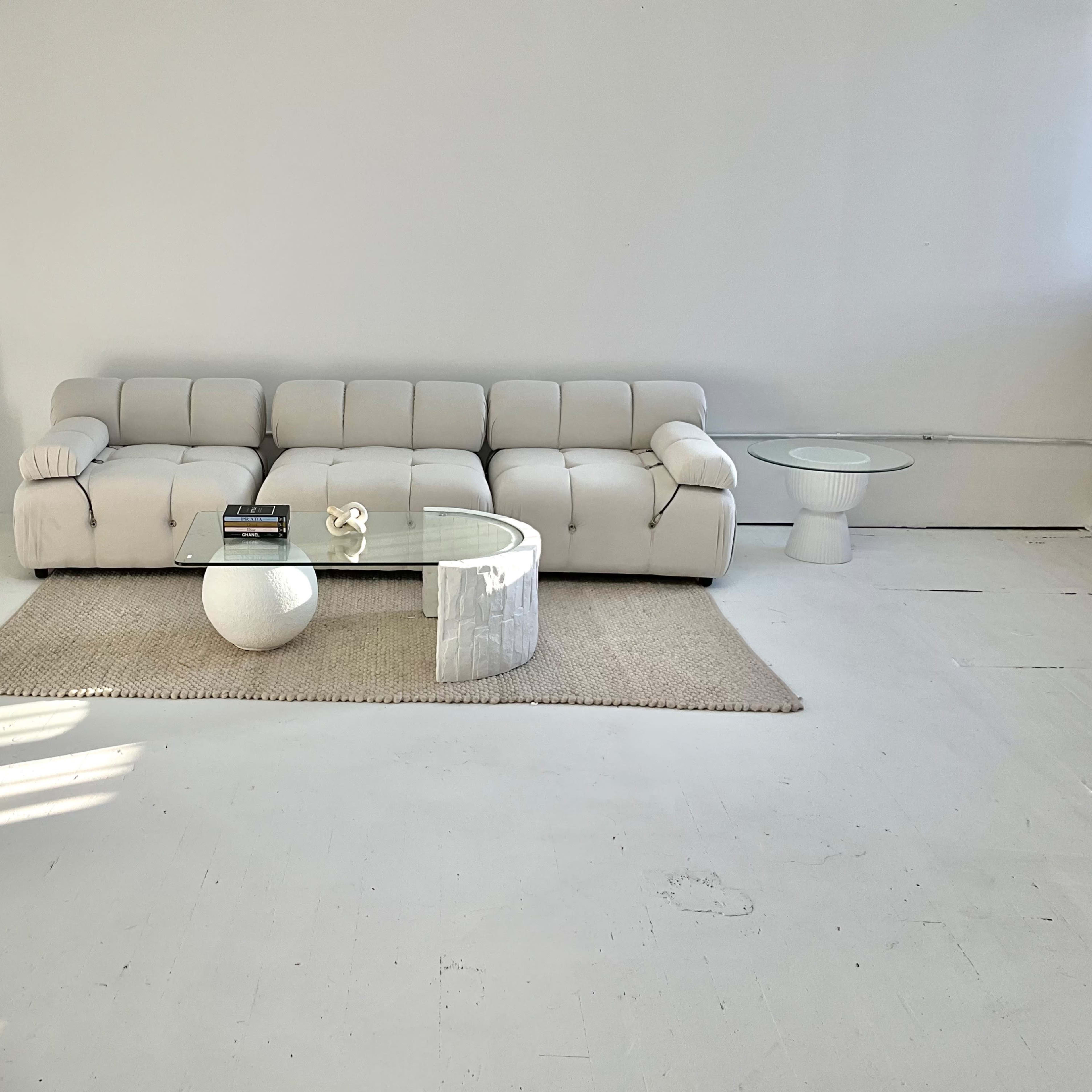 A light-colored sectional sofa is placed on a woven rug in a minimalist room, accompanied by a glass coffee table and a small white side table.