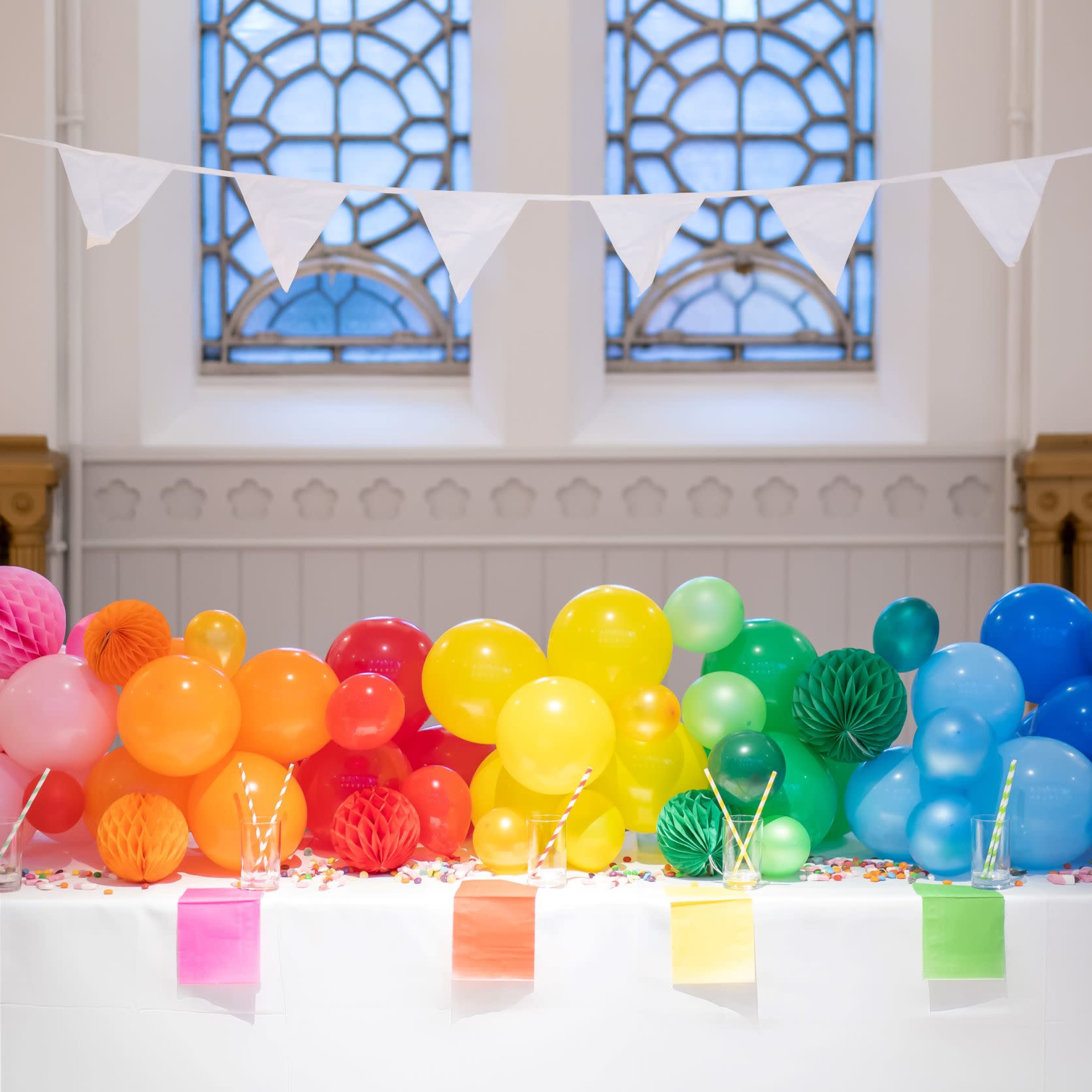 A table decorated with brightly colored balloons and festive paper decorations is set against a backdrop of large, stained glass windows.