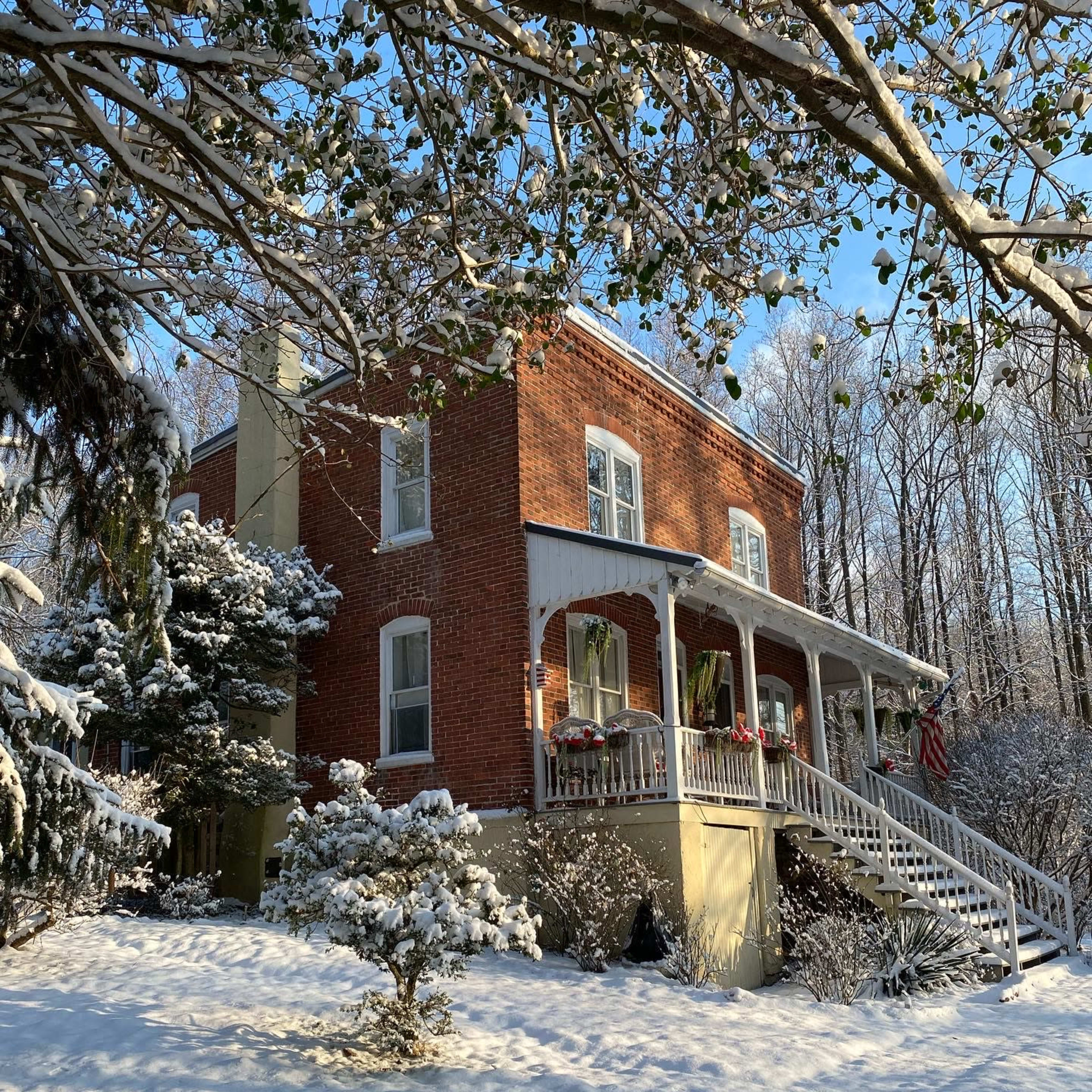 A two-story brick house with a front porch is surrounded by snow-covered trees and bushes on a sunny winter day.