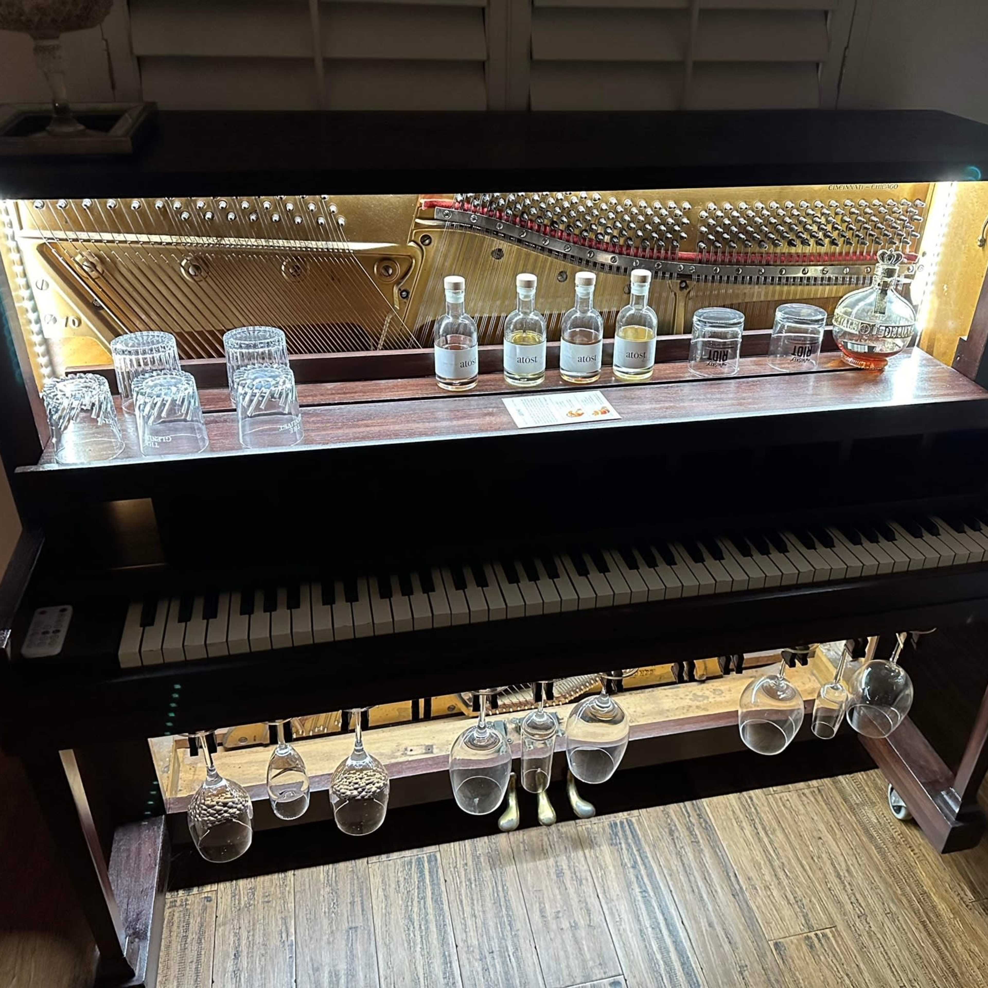 A vintage piano is converted into a bar display, holding several bottles and glassware illuminated from above.