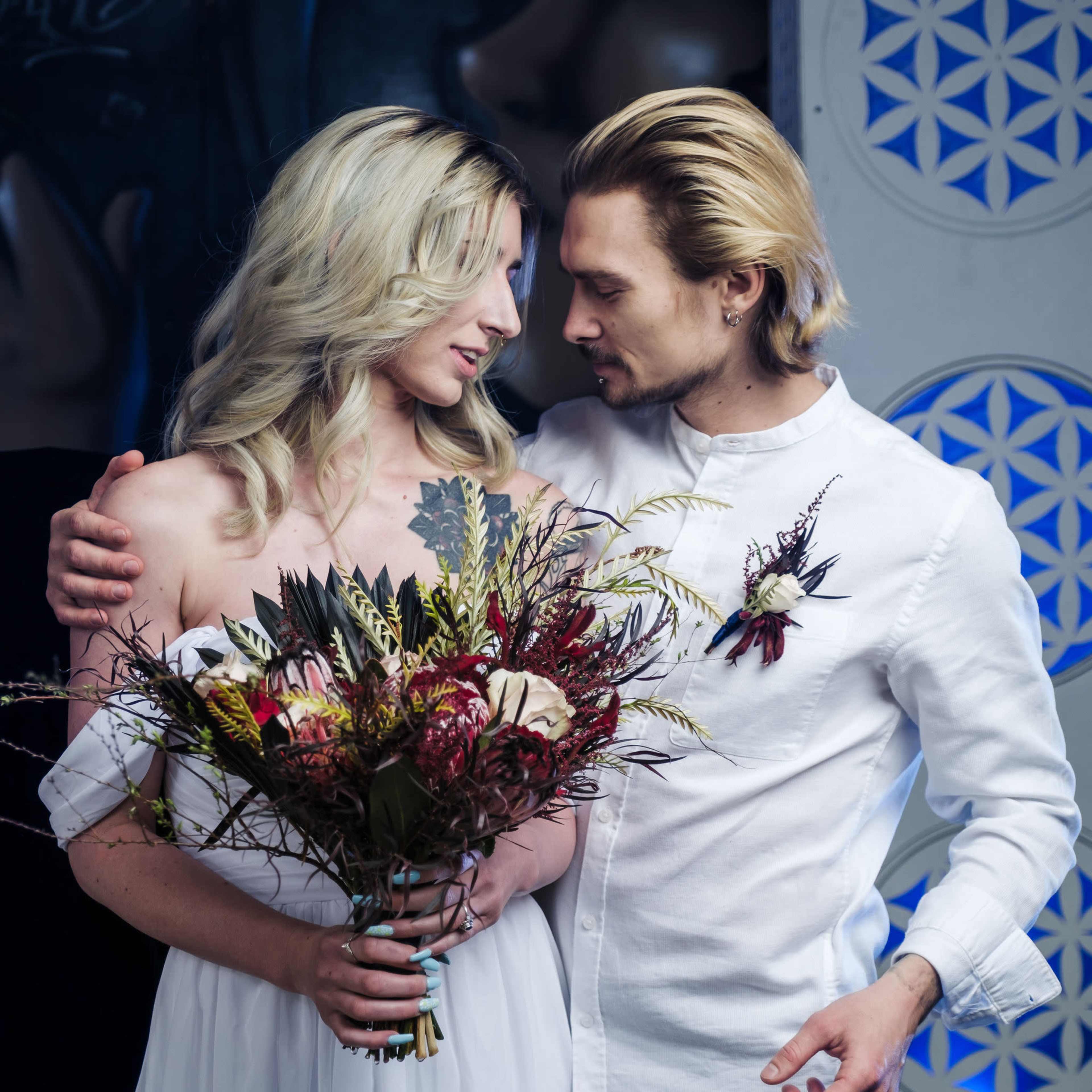 A couple stands close together, with the woman holding a bouquet of dried flowers while gazing at the man, who is dressed in a white shirt.