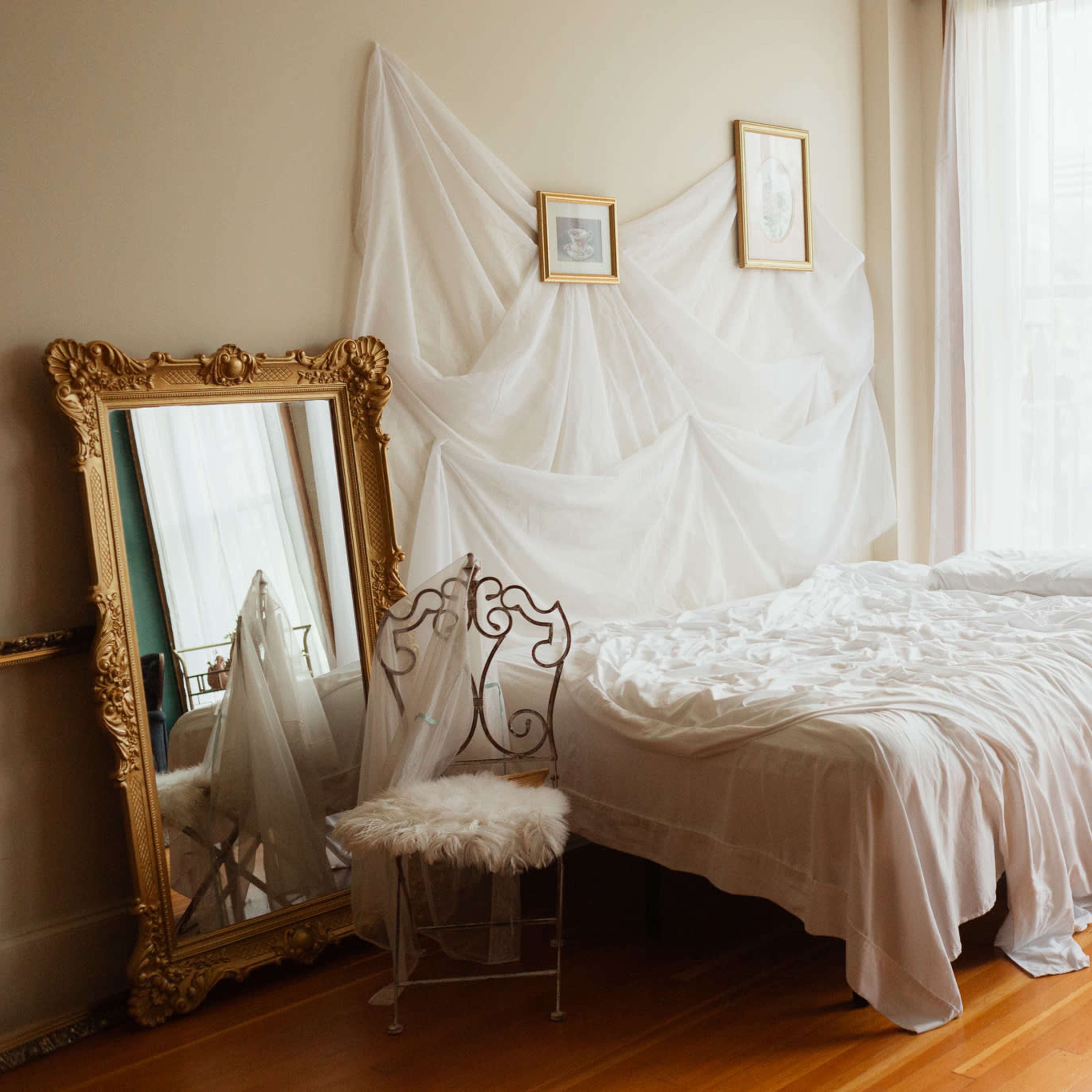 A neatly made bed with white linens is positioned next to a white nightstand and a tall mirror in a bright room adorned with sheer drapes.
