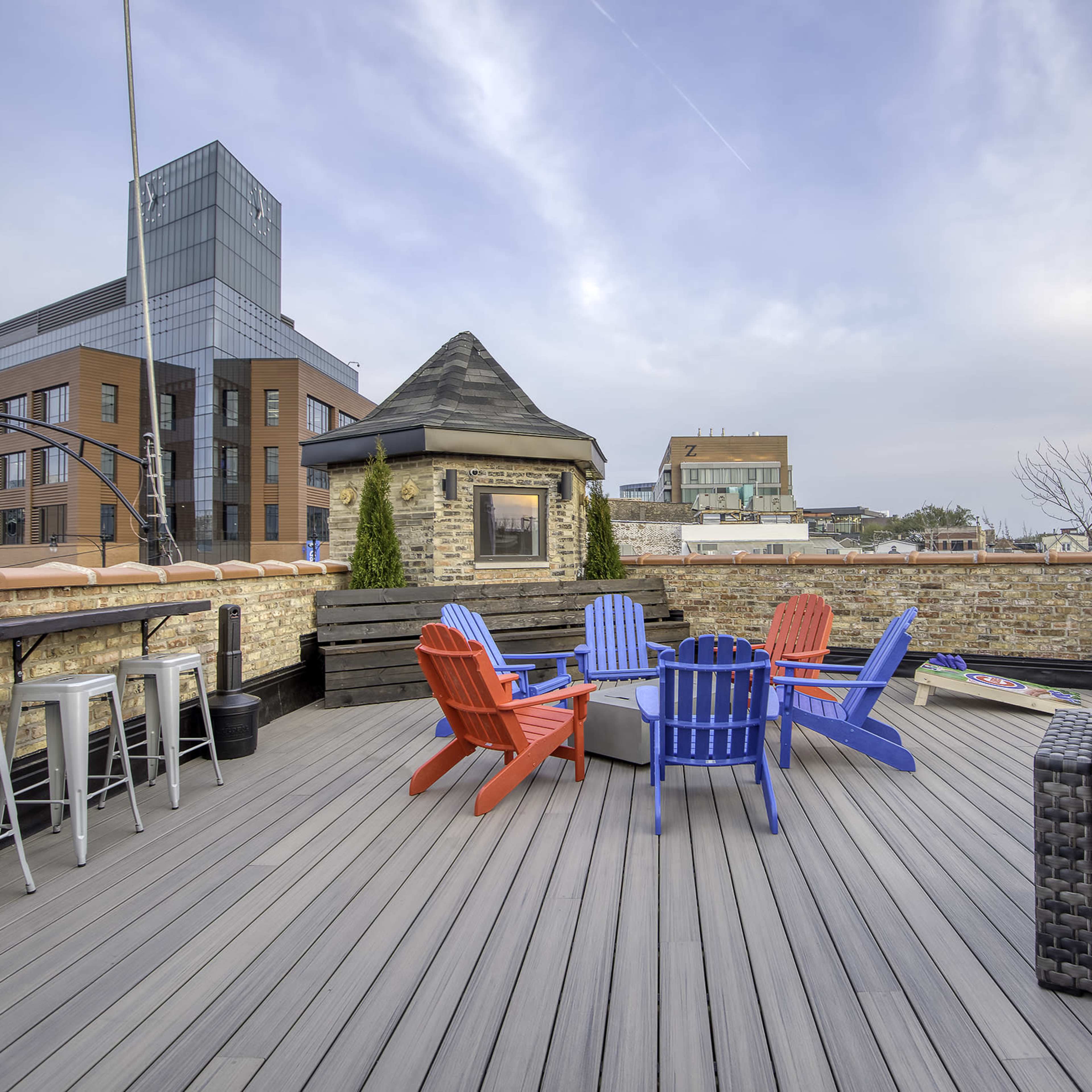 A rooftop deck with colorful adirondack chairs, a seating area, and a bar with high stools, surrounded by city buildings.