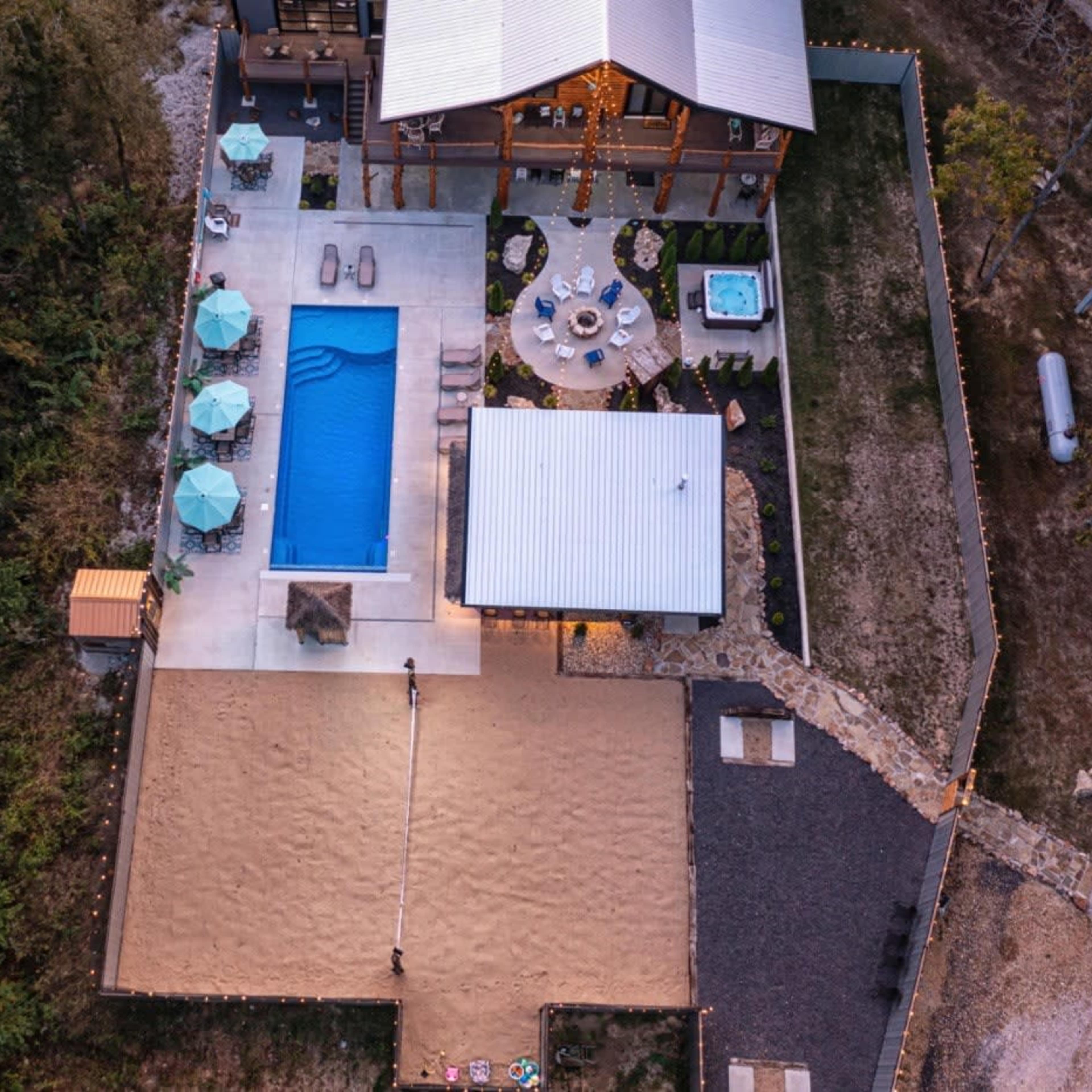 An aerial view of a property featuring a swimming pool, a hot tub, outdoor seating areas, and a sand volleyball court, surrounded by trees.