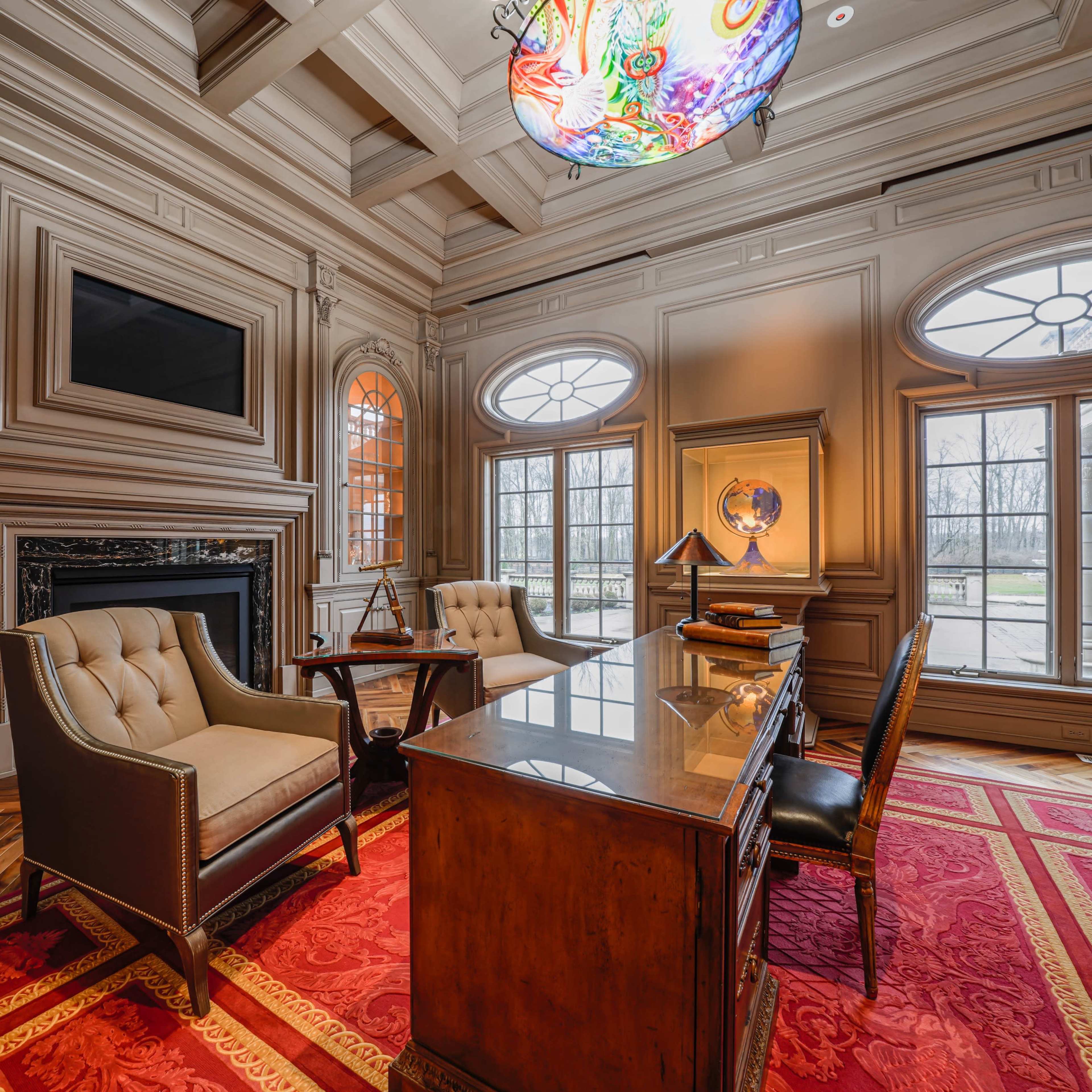An elegantly furnished office featuring a wooden desk, two armchairs, large windows, and ornate wall detailing.