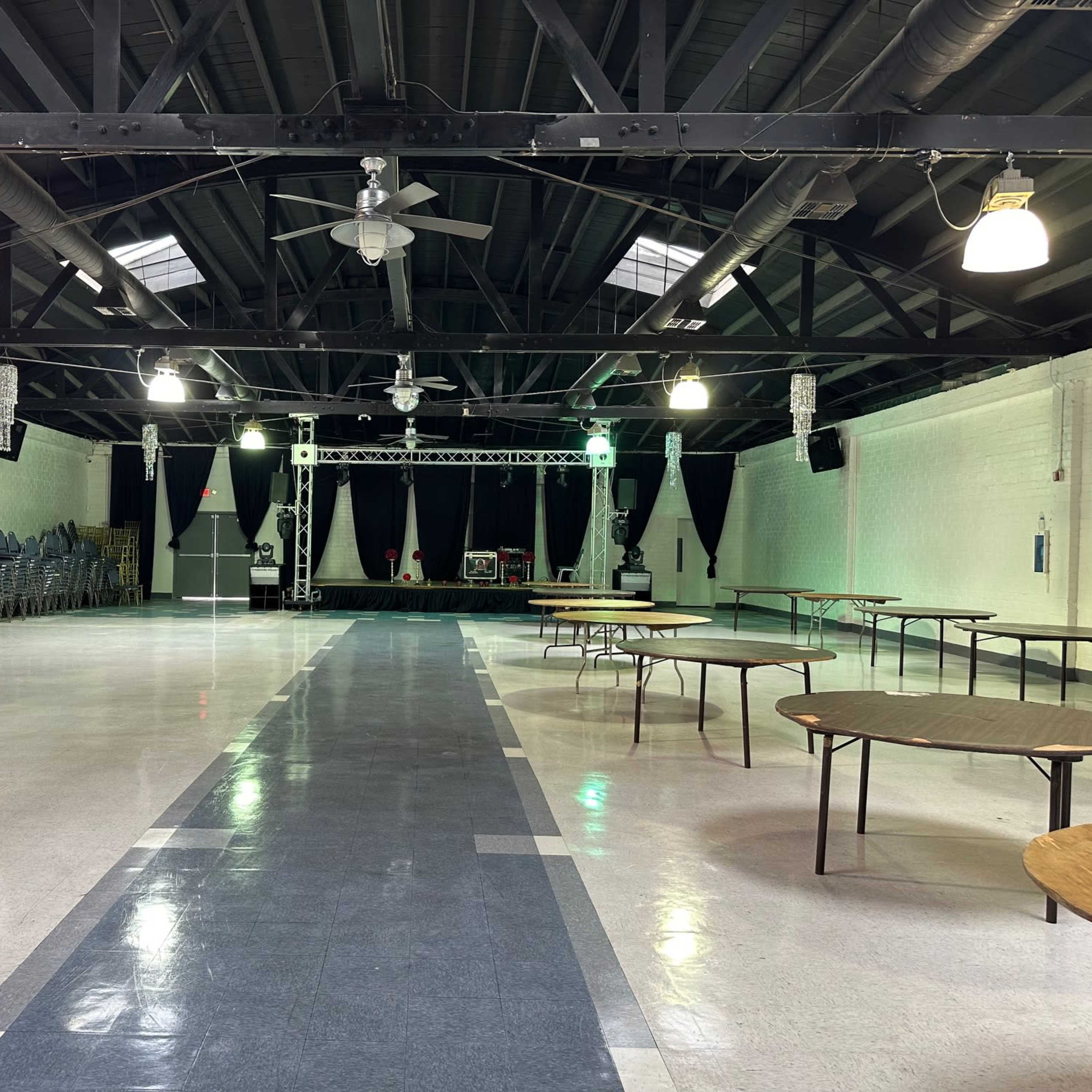 A large, vacant indoor hall features rows of stacked chairs on one side and several empty tables arranged throughout the space, with a stage at the far end.