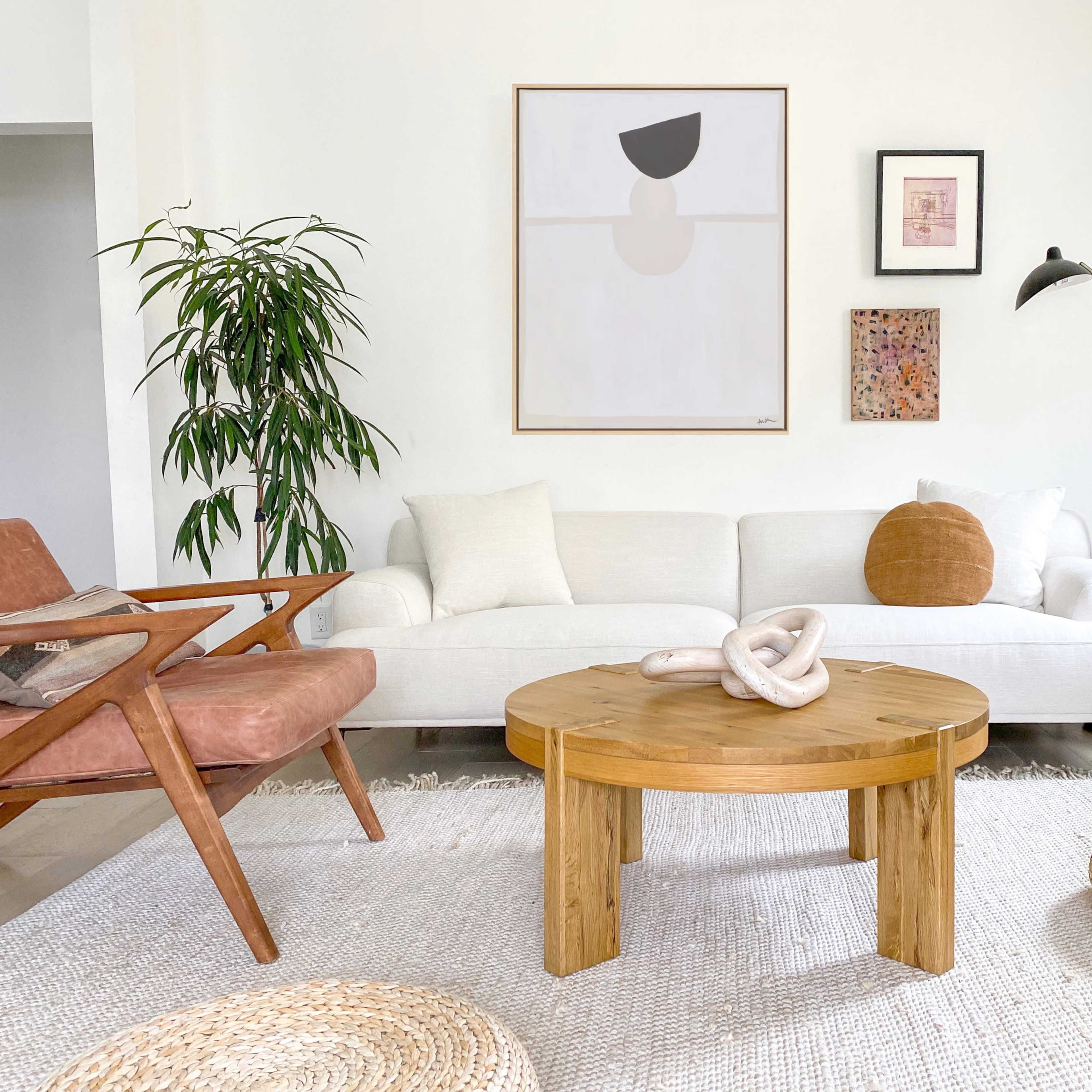 A bright living room features a white couch, a round wooden coffee table, a large rug, and various decorative elements, including plants and artwork on the walls.