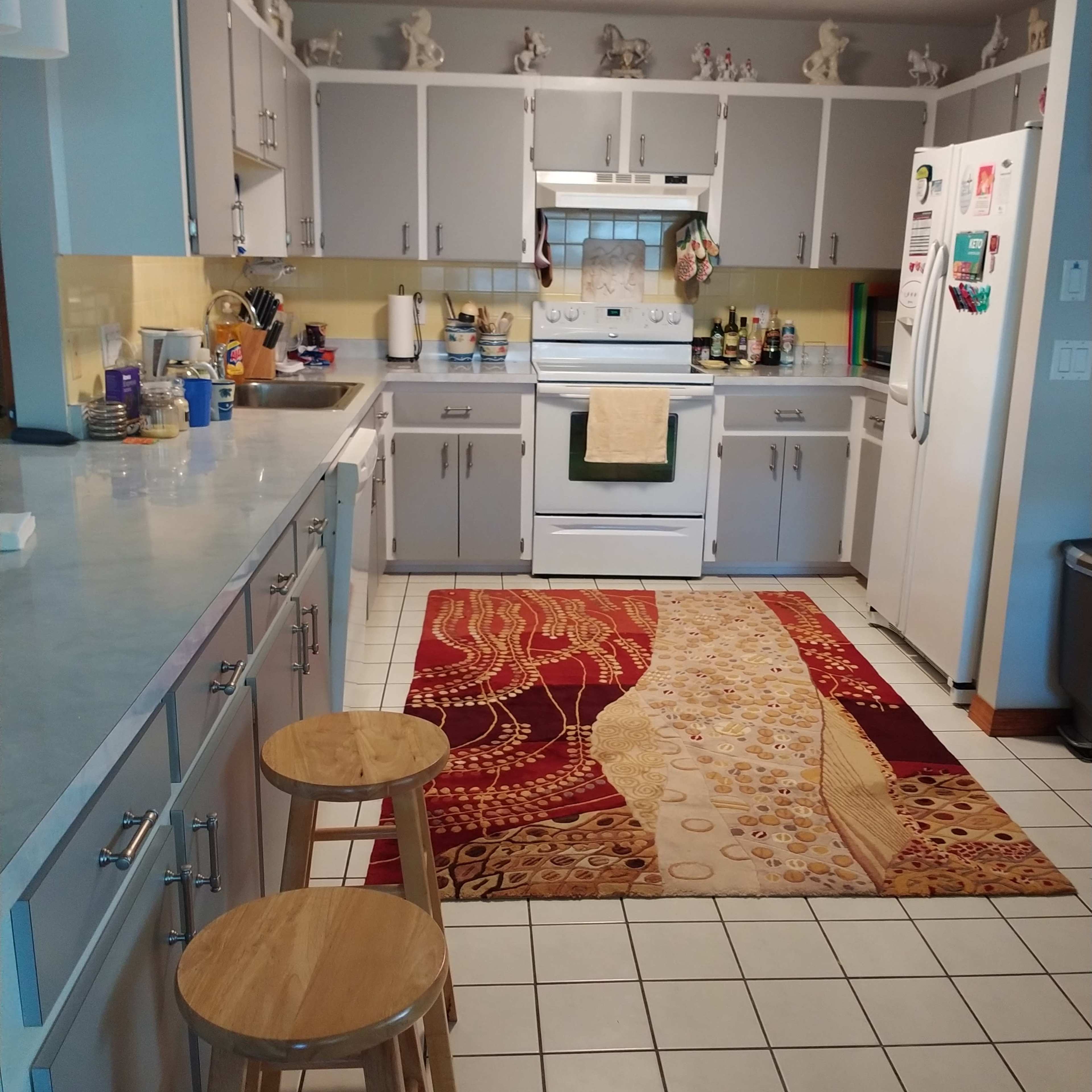 A kitchen featuring light gray cabinets, a white stove and refrigerator, and a patterned area rug on the tiled floor.