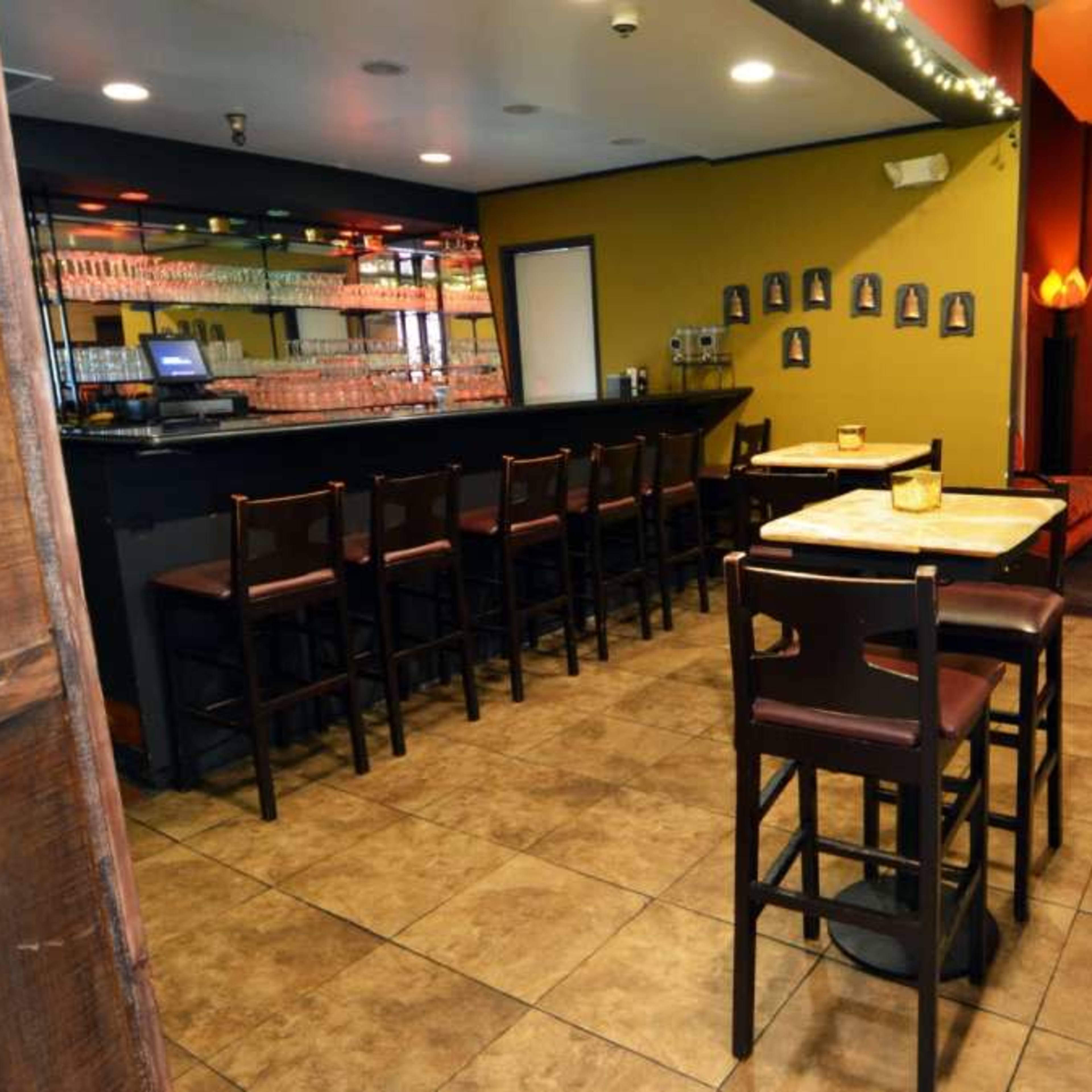 The interior of a bar featuring a long counter with bar stools, several tables, and walls adorned with small framed pictures.