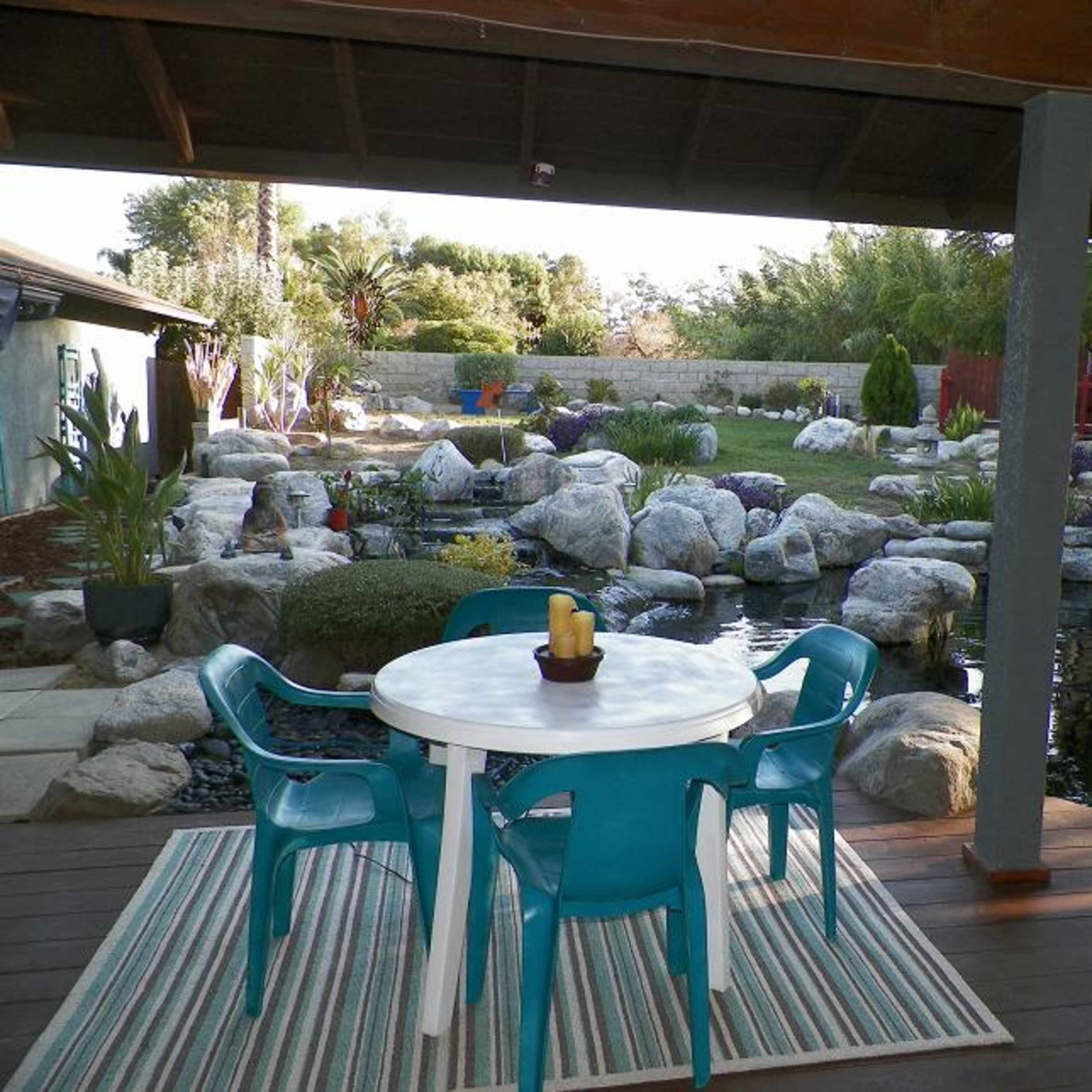 A dining table with blue chairs sits on a wooden patio overlooking a landscaped yard with a pond and rock features.