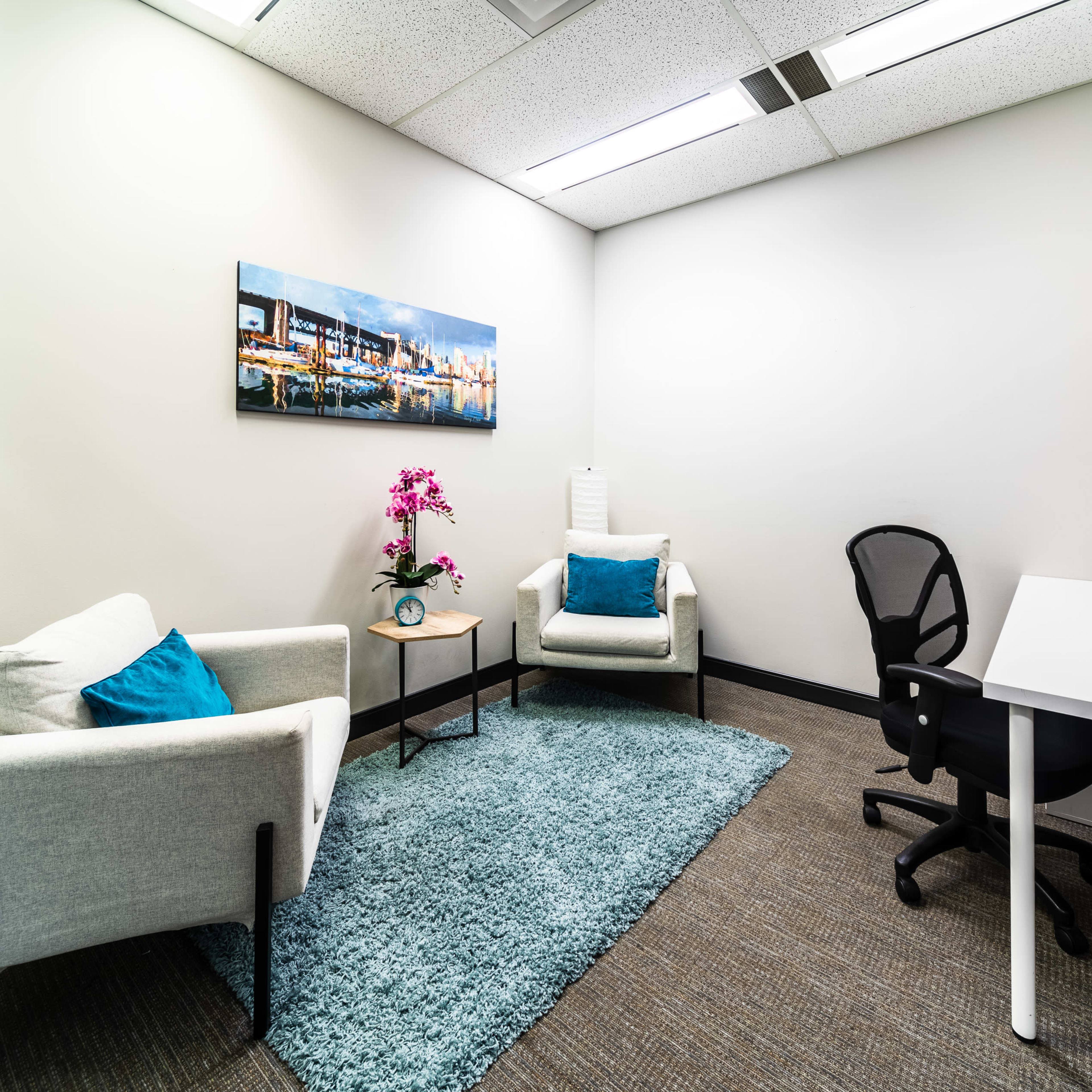 A small office space featuring two armchairs, a desk with a chair, a rug, and decorative plants, along with a painting on the wall.