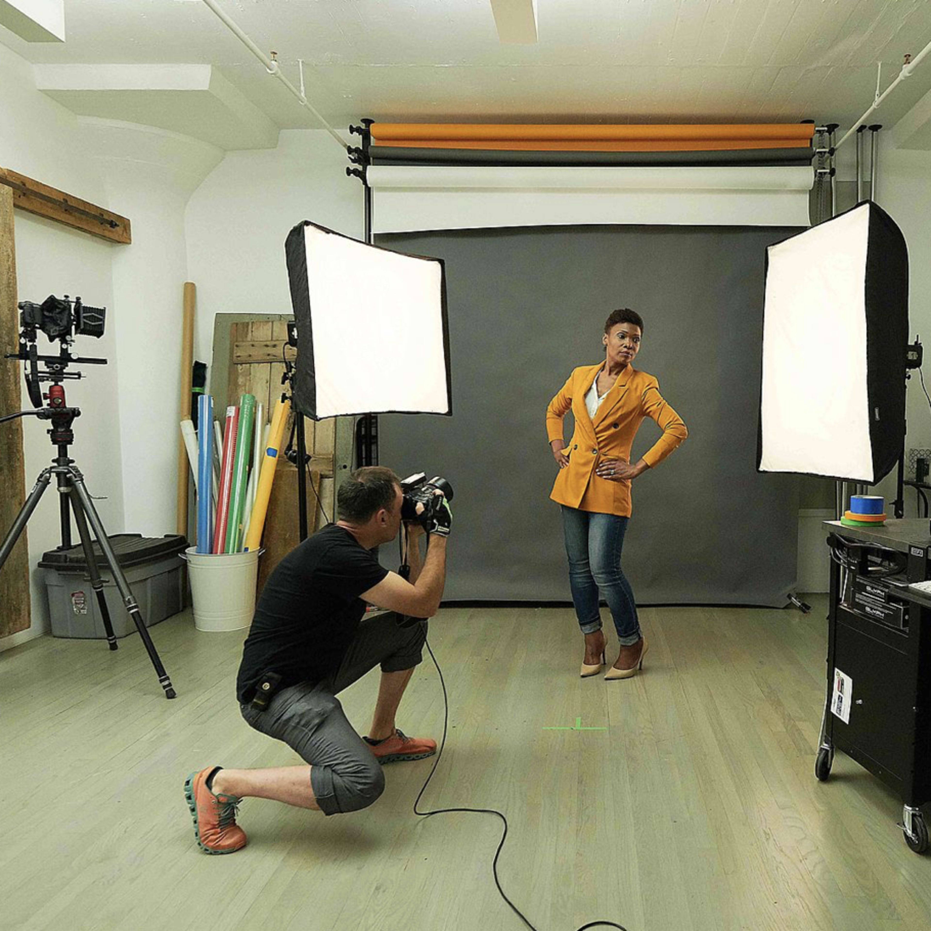 A photographer kneels to take a picture of a model posing in front of a backdrop in a photography studio.