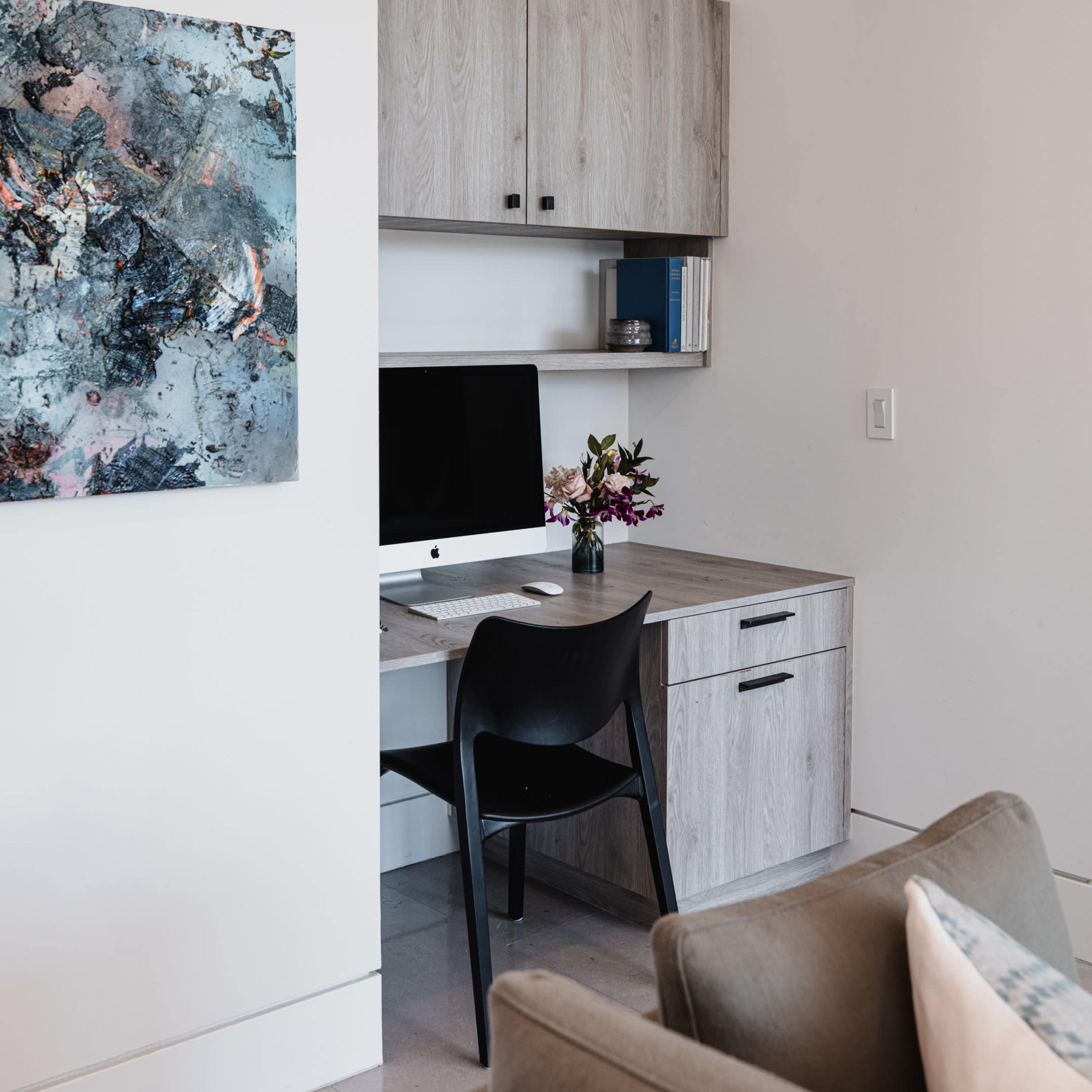 A modern home office features a desk with an Apple computer, surrounded by light wood cabinetry and a decorative painting on the wall.