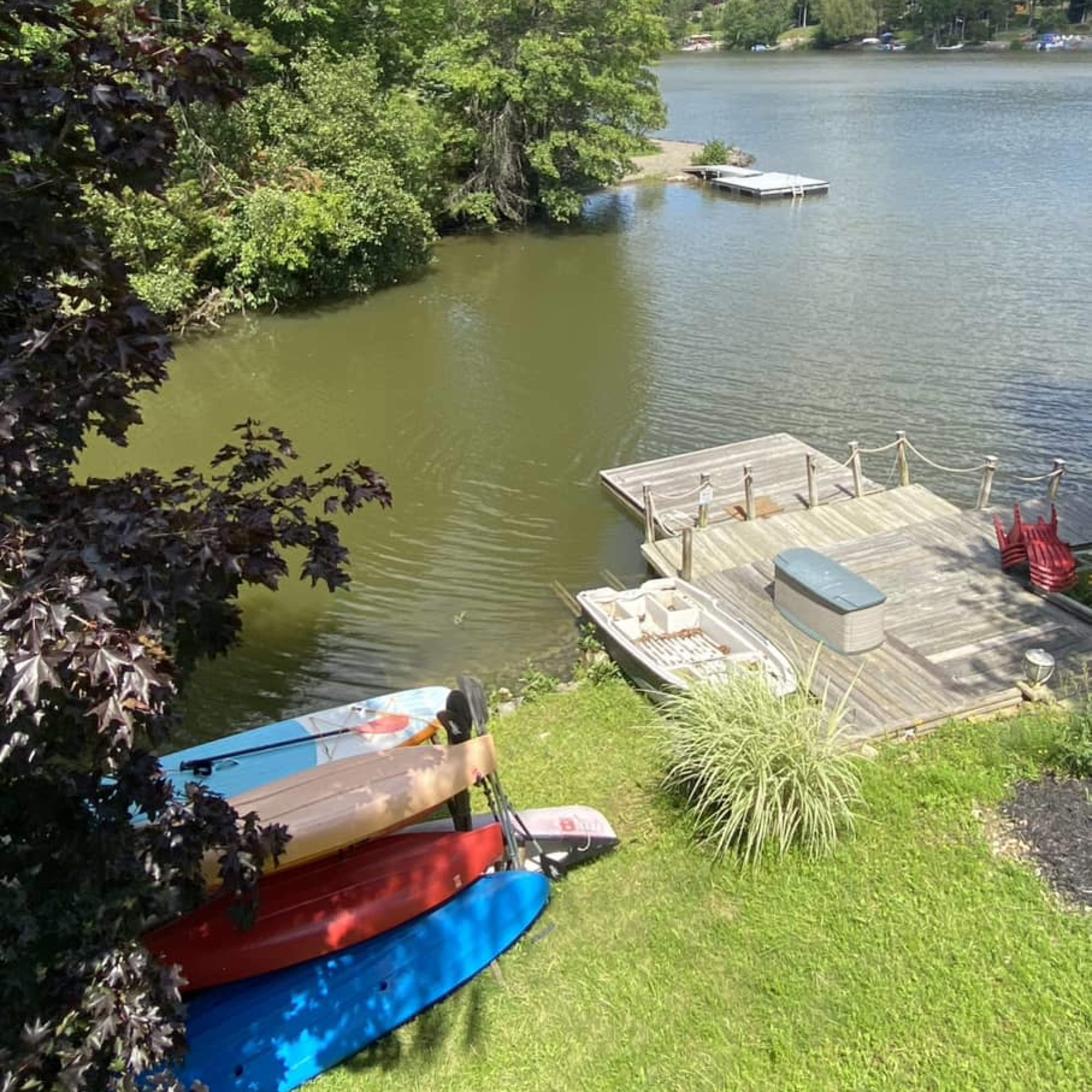 A collection of colorful kayaks and canoes are lined up on the grass near a calm lake with a wooden dock in the background.