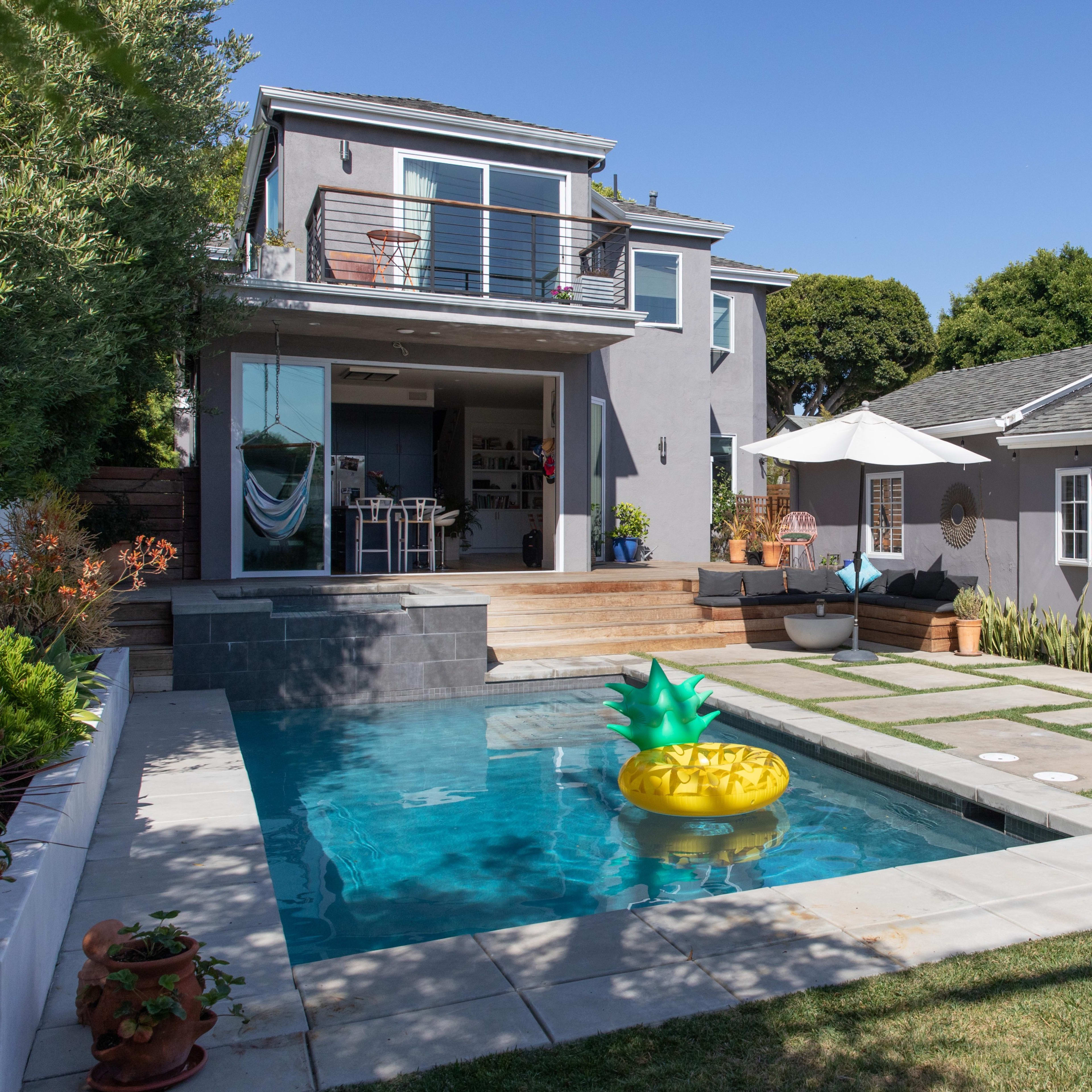 A modern backyard features a swimming pool with a pineapple-shaped float, surrounded by landscaped grass and two adjoining structures.