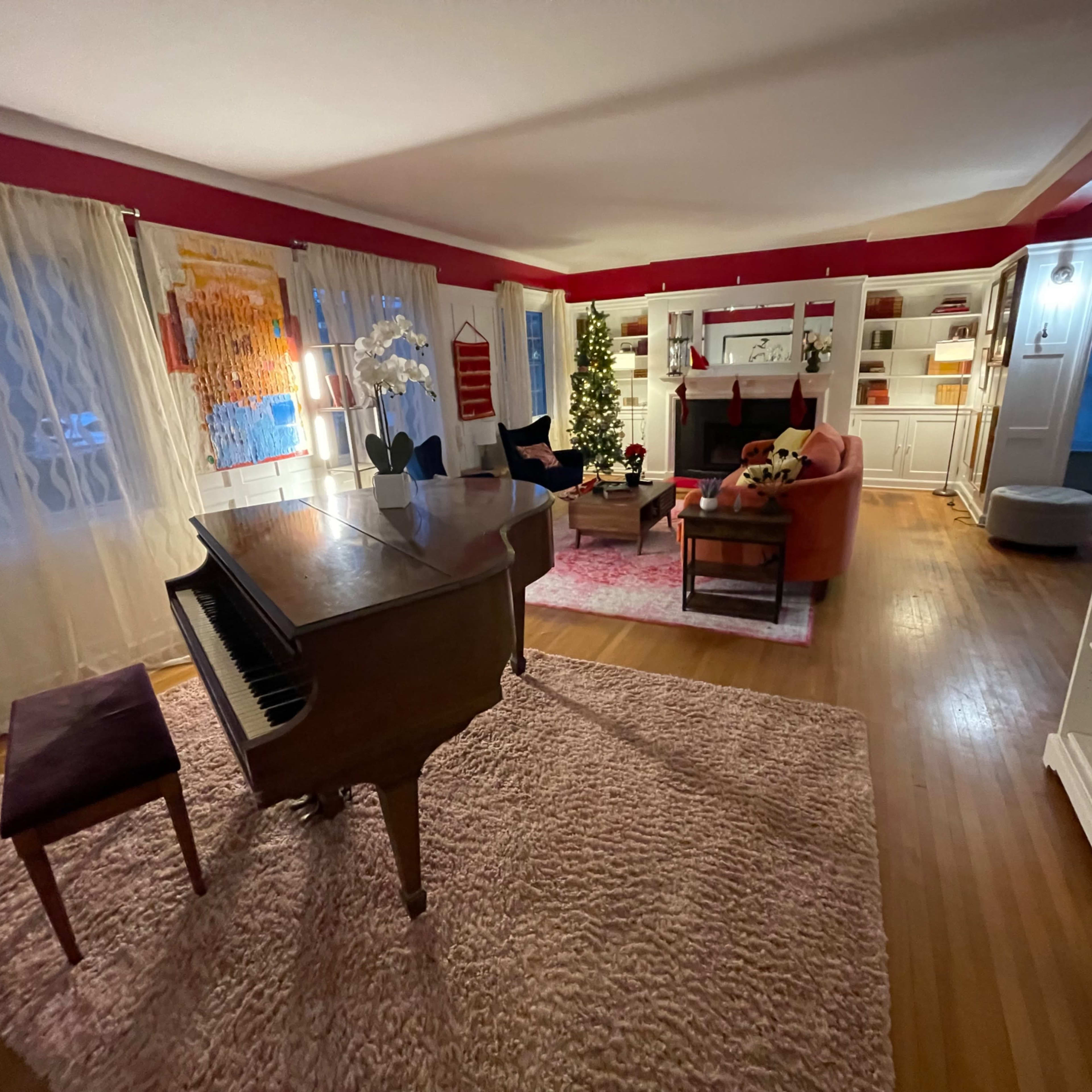 A spacious living room with a grand piano, a large area rug, and decorated shelves, featuring a Christmas tree and a cozy seating arrangement.