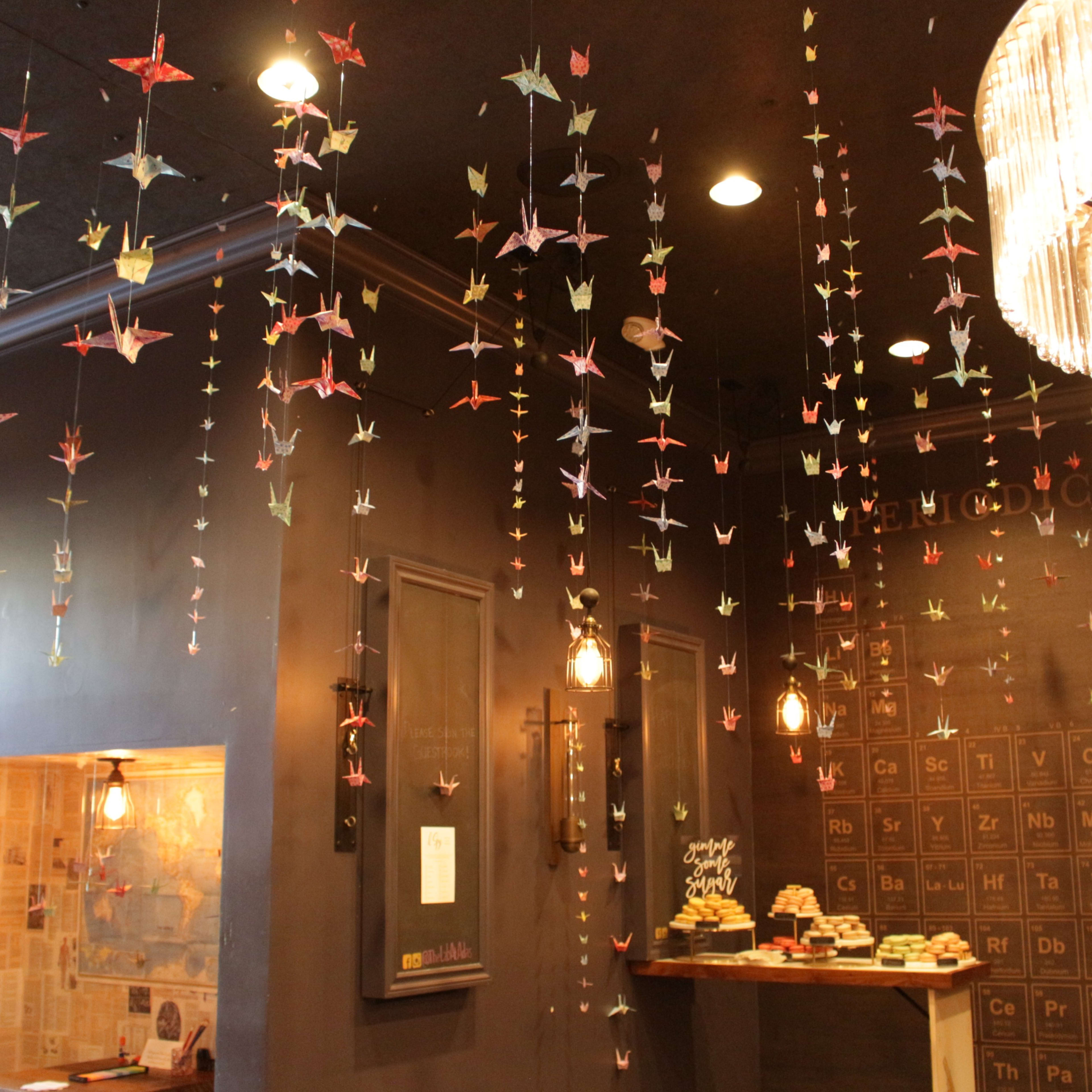 The interior of a café features many hanging paper cranes and a large chandelier above a counter with pastries.