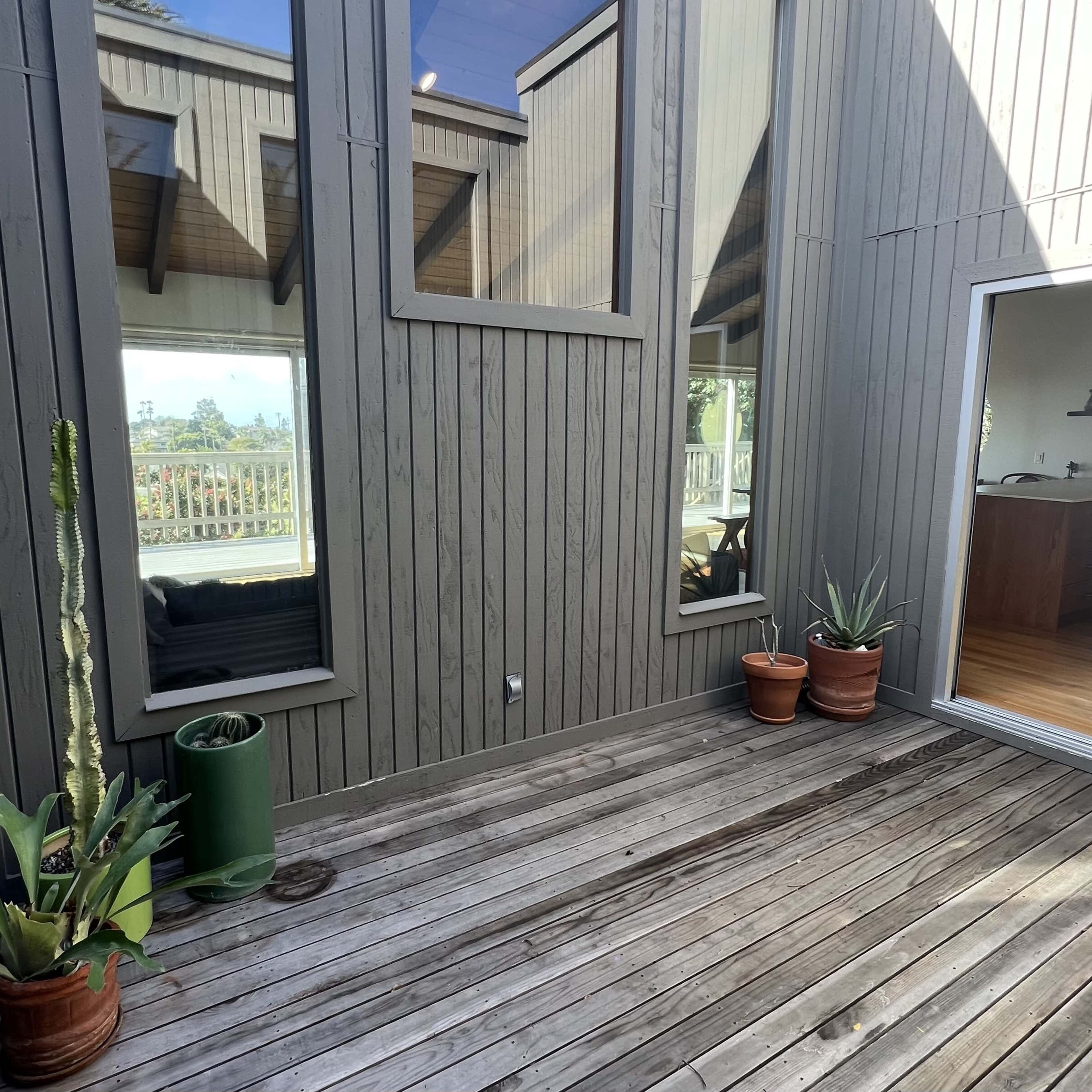 A wooden deck area surrounded by gray vertical walls with large windows reflecting the exterior landscape and containing potted plants.