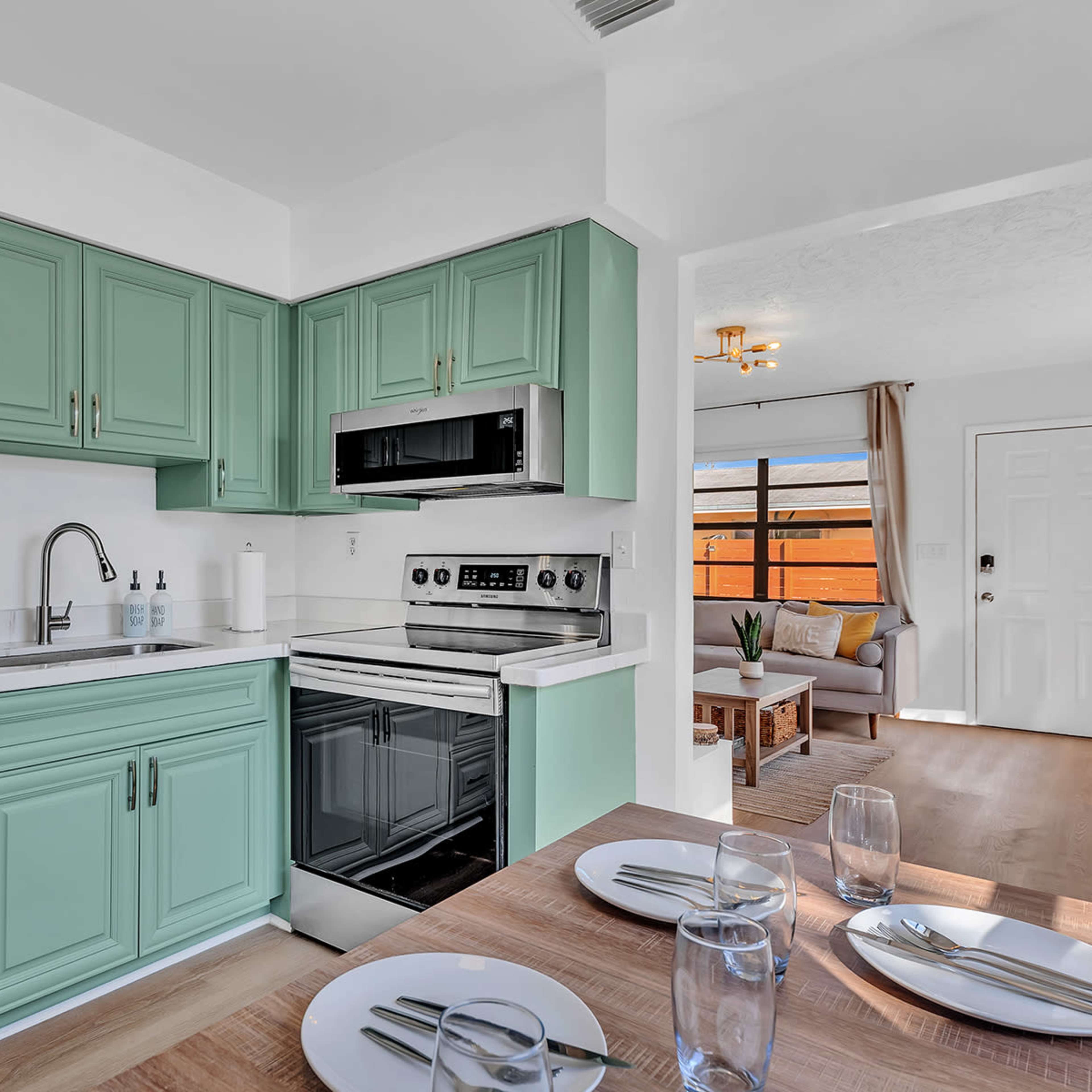 A modern kitchen with mint green cabinets and stainless steel appliances, adjacent to a dining area with a table set for four.