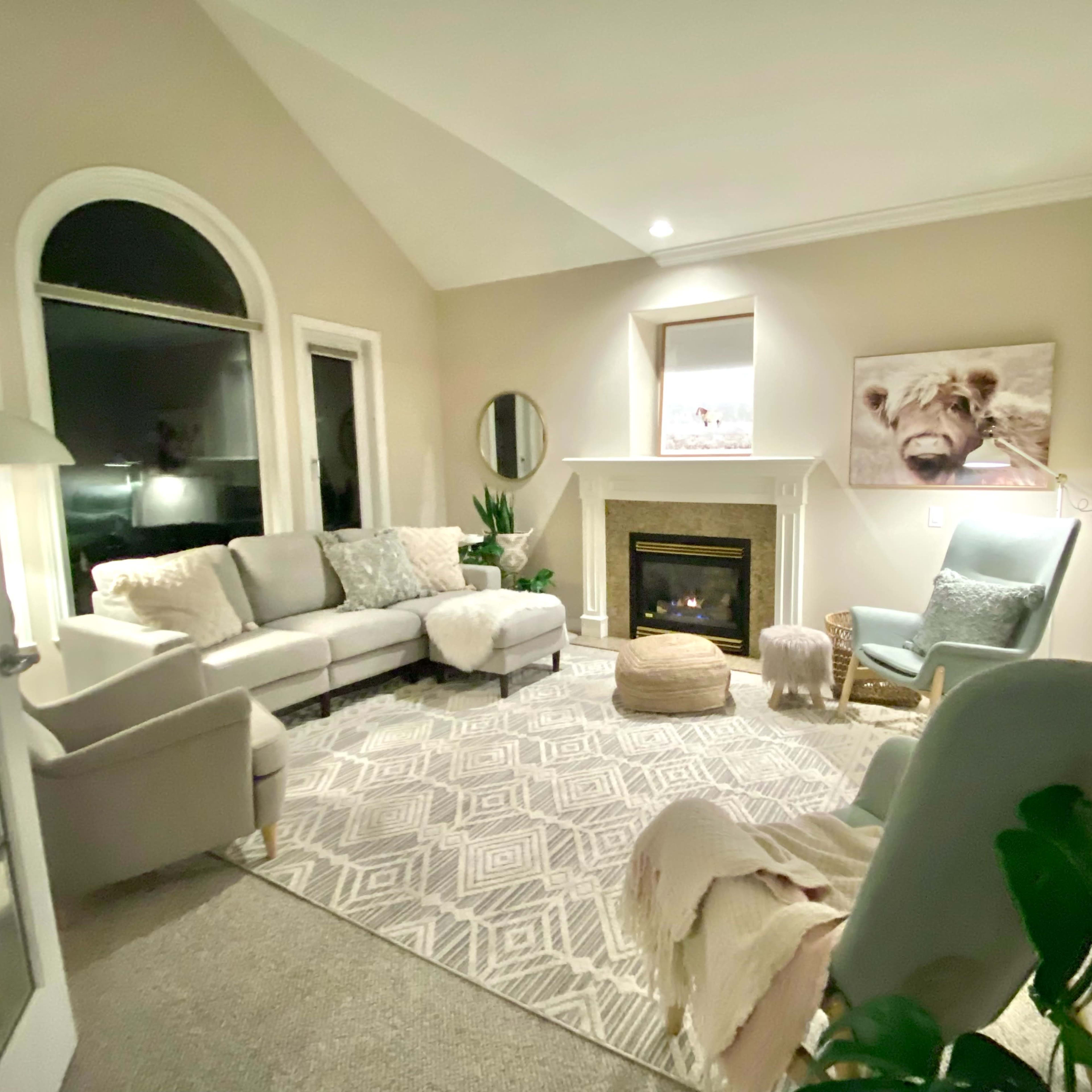 A cozy living room with a fireplace, a large area rug, and comfortable seating arrangements, including a light gray sofa and a green armchair.