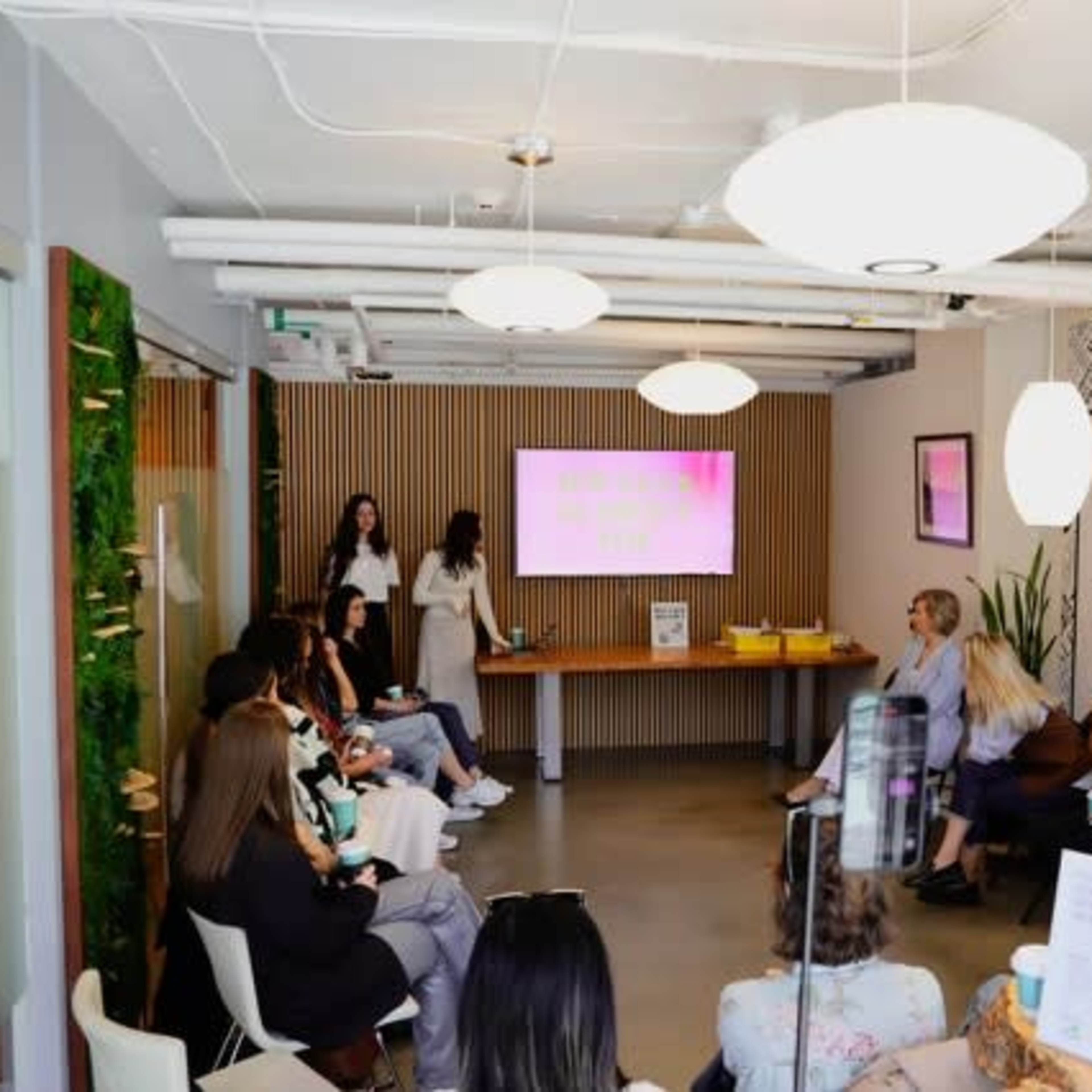 A group of people sits in a modern conference room, attentively watching a presentation on a screen while two presenters stand at the front.
