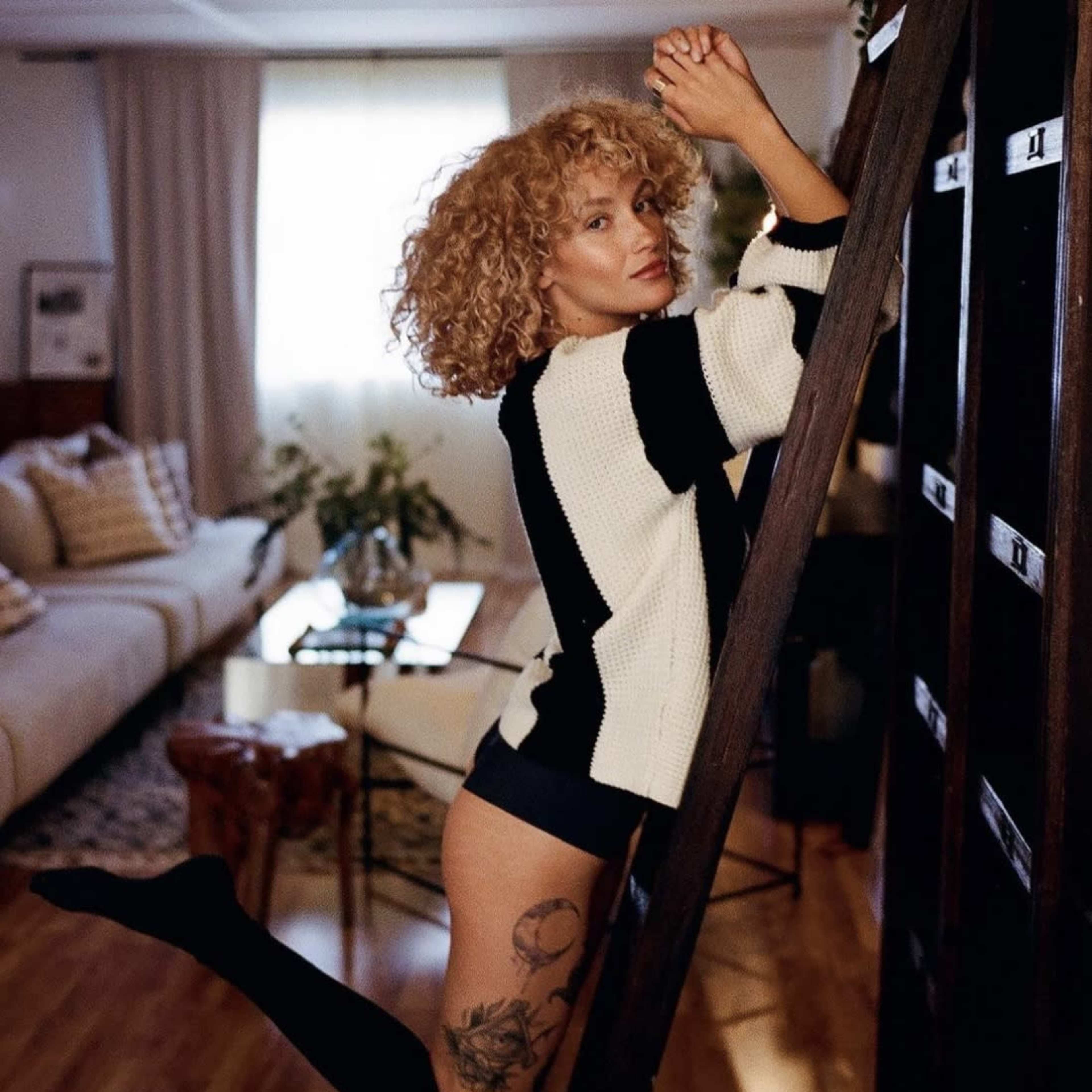 A person with curly hair poses on a wooden ladder in a well-lit room, showcasing a tattoo on their leg.