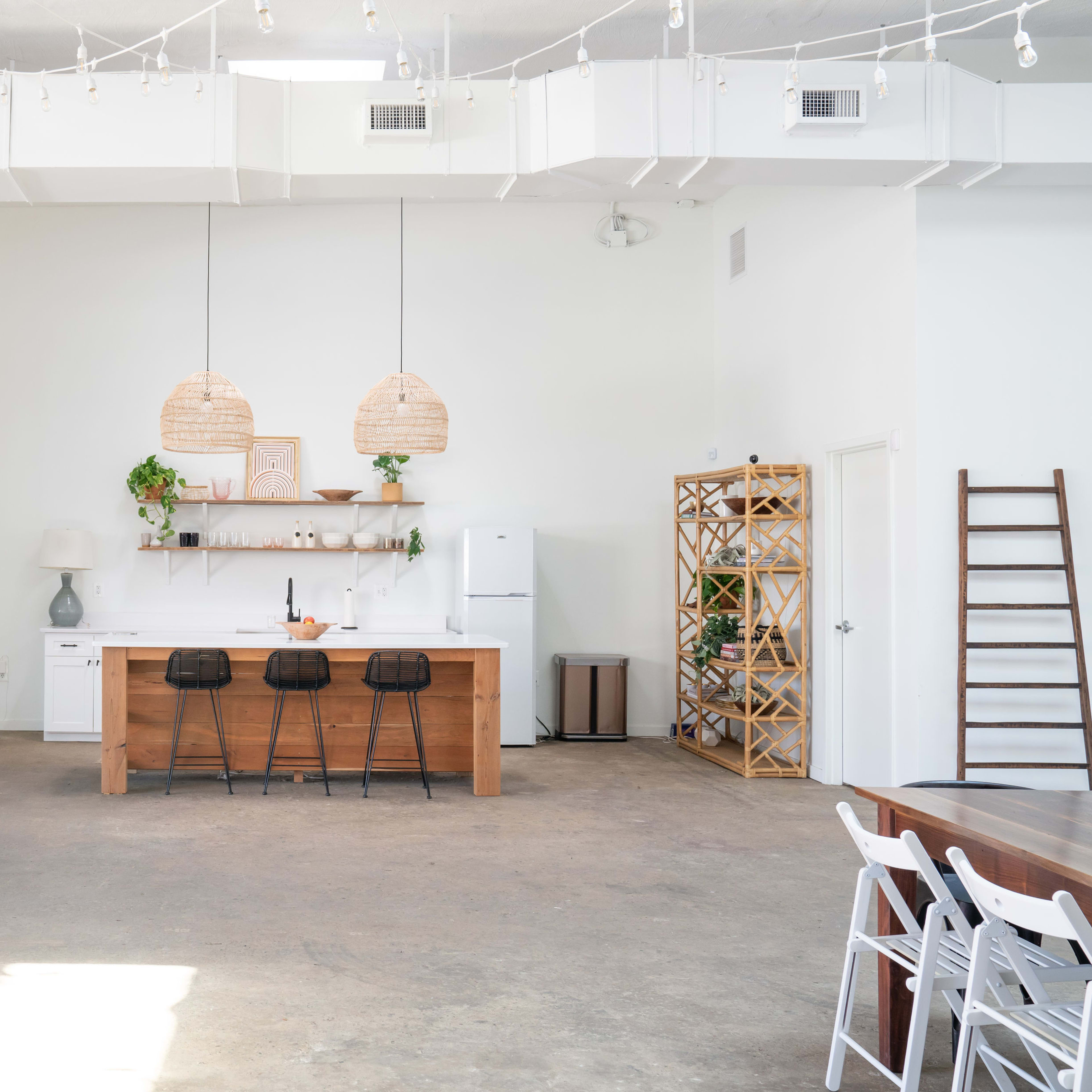 A spacious, modern kitchen with a wooden island, three black bar stools, and pendant lighting above, complemented by open shelving and a refrigerator in a light-filled environment.