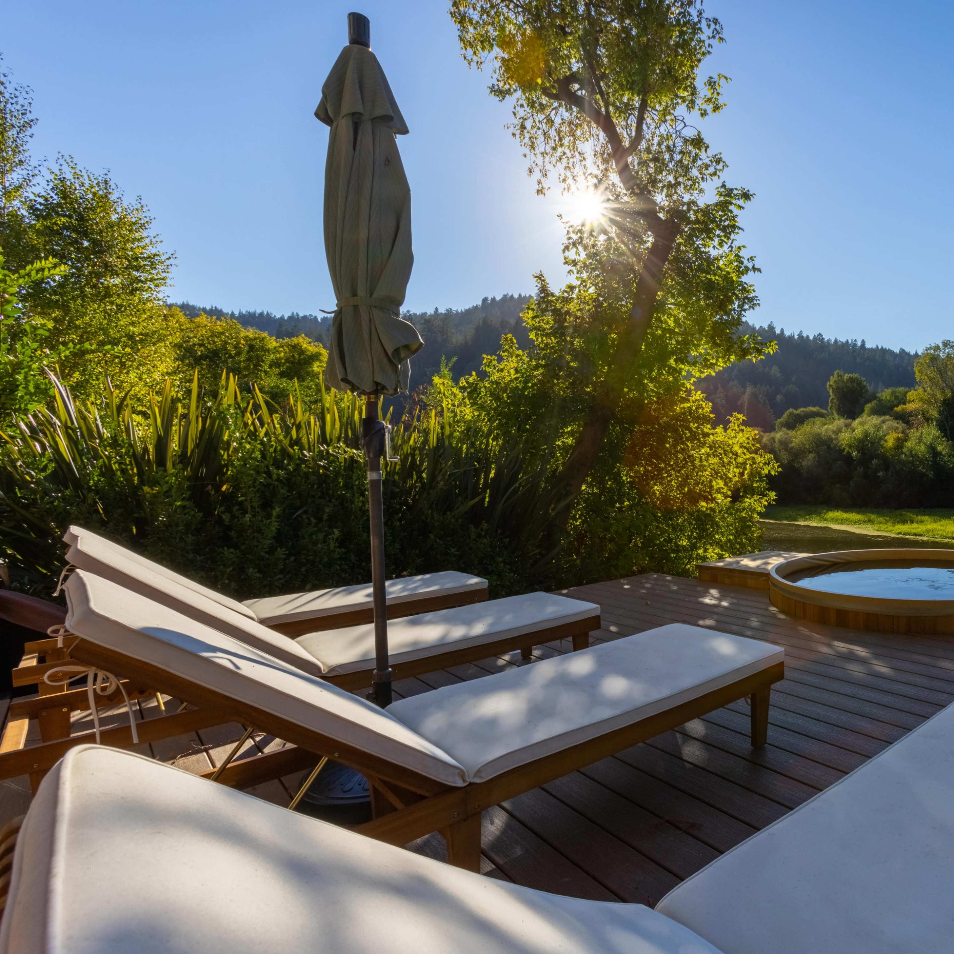 A wooden deck with lounge chairs and a round hot tub overlooks a lush green area surrounded by trees under a clear blue sky.