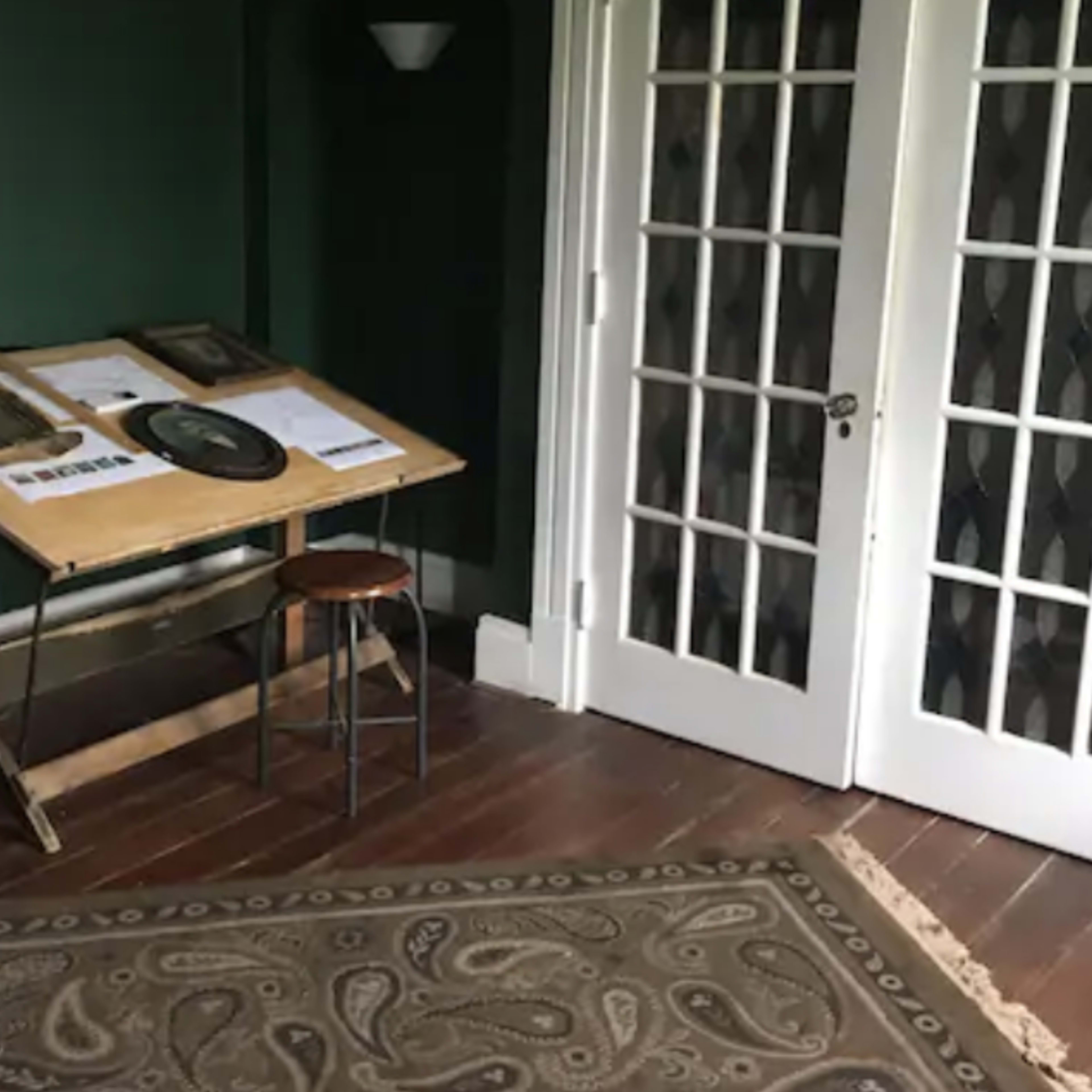 A wooden table with a stool is positioned near a set of double doors in a room featuring dark green walls and a patterned rug on the wooden floor.