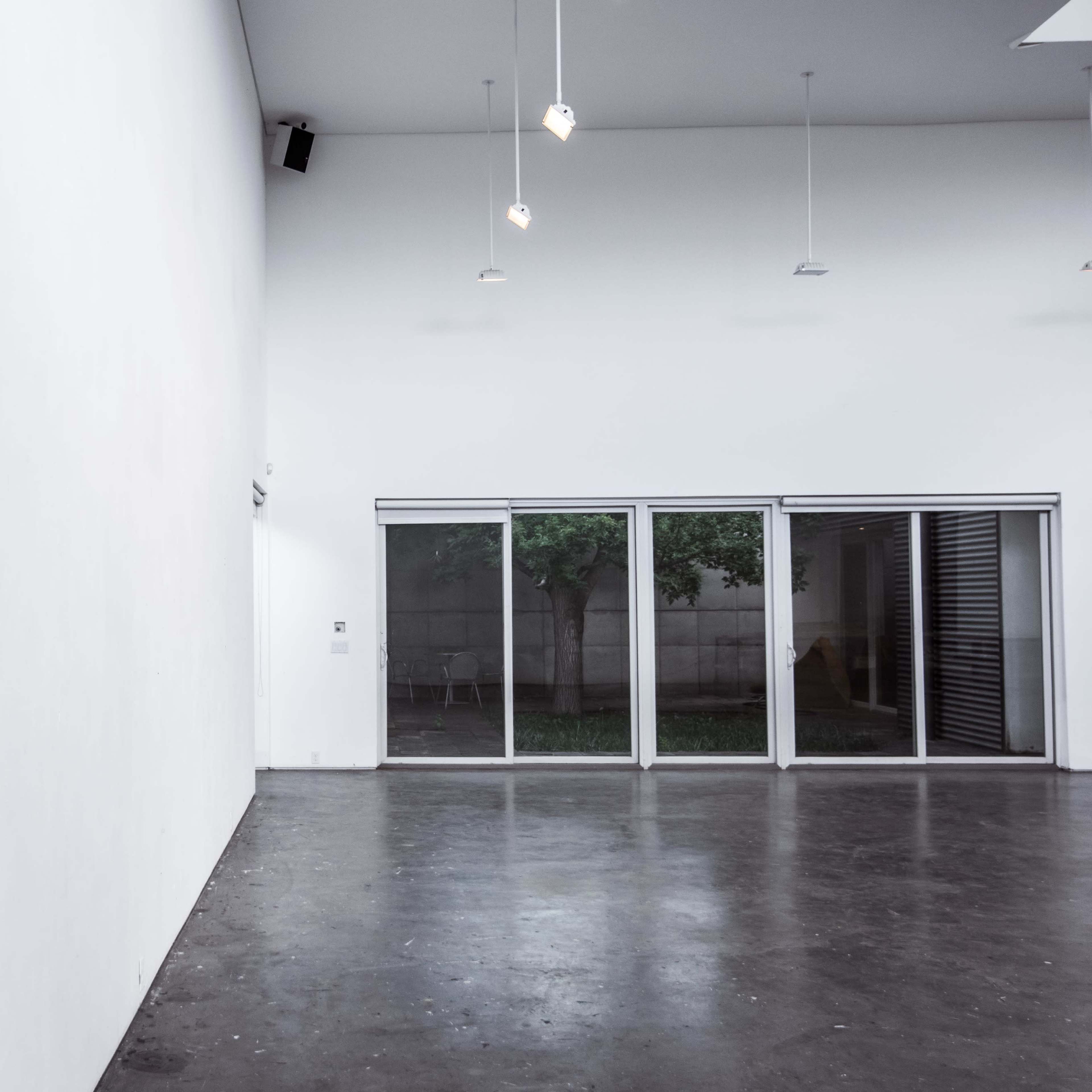 A minimalist interior space with a large white wall and sliding glass doors leading to an outdoor area with visible greenery.