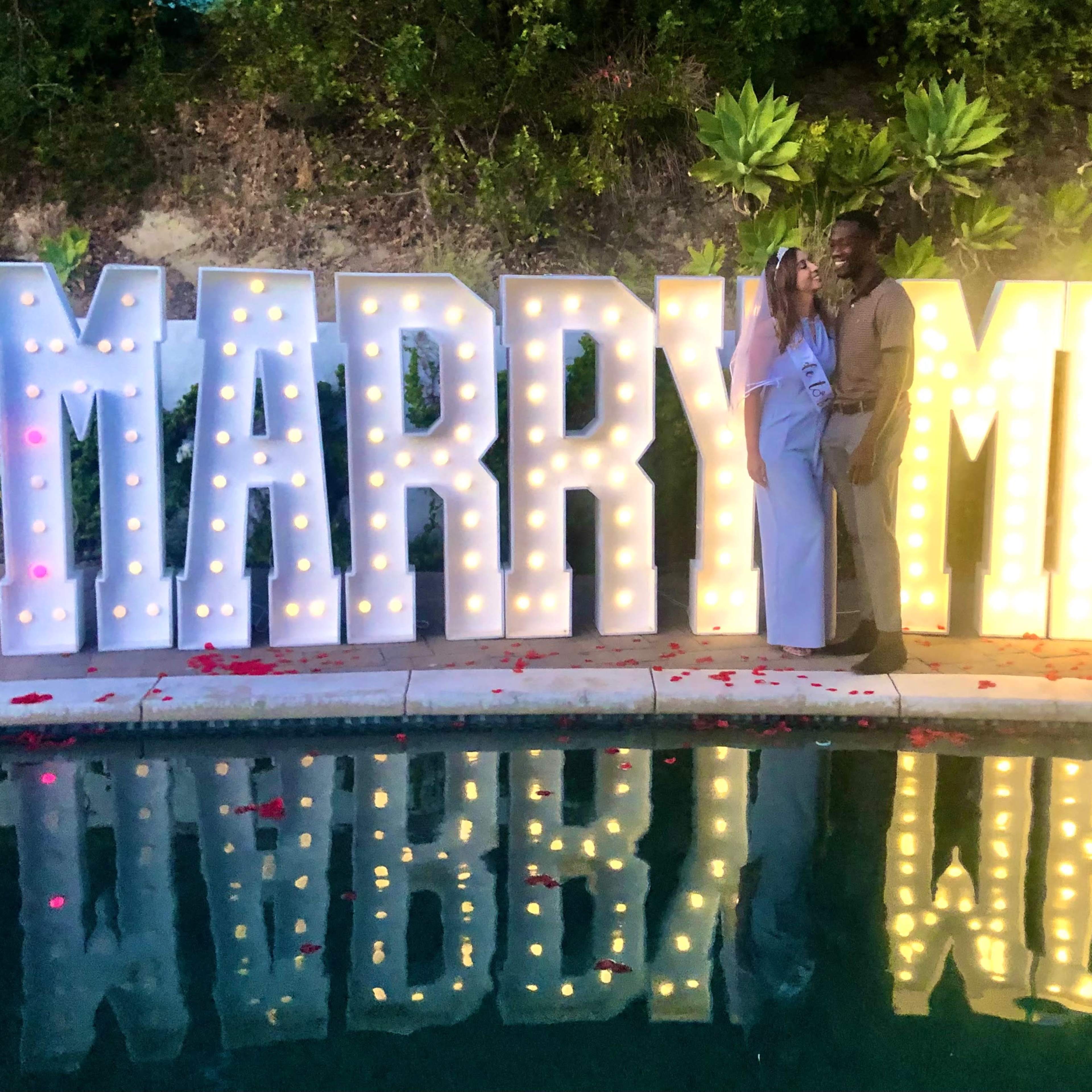 A couple poses in front of illuminated "MARRY ME" letters near a pool surrounded by rose petals.