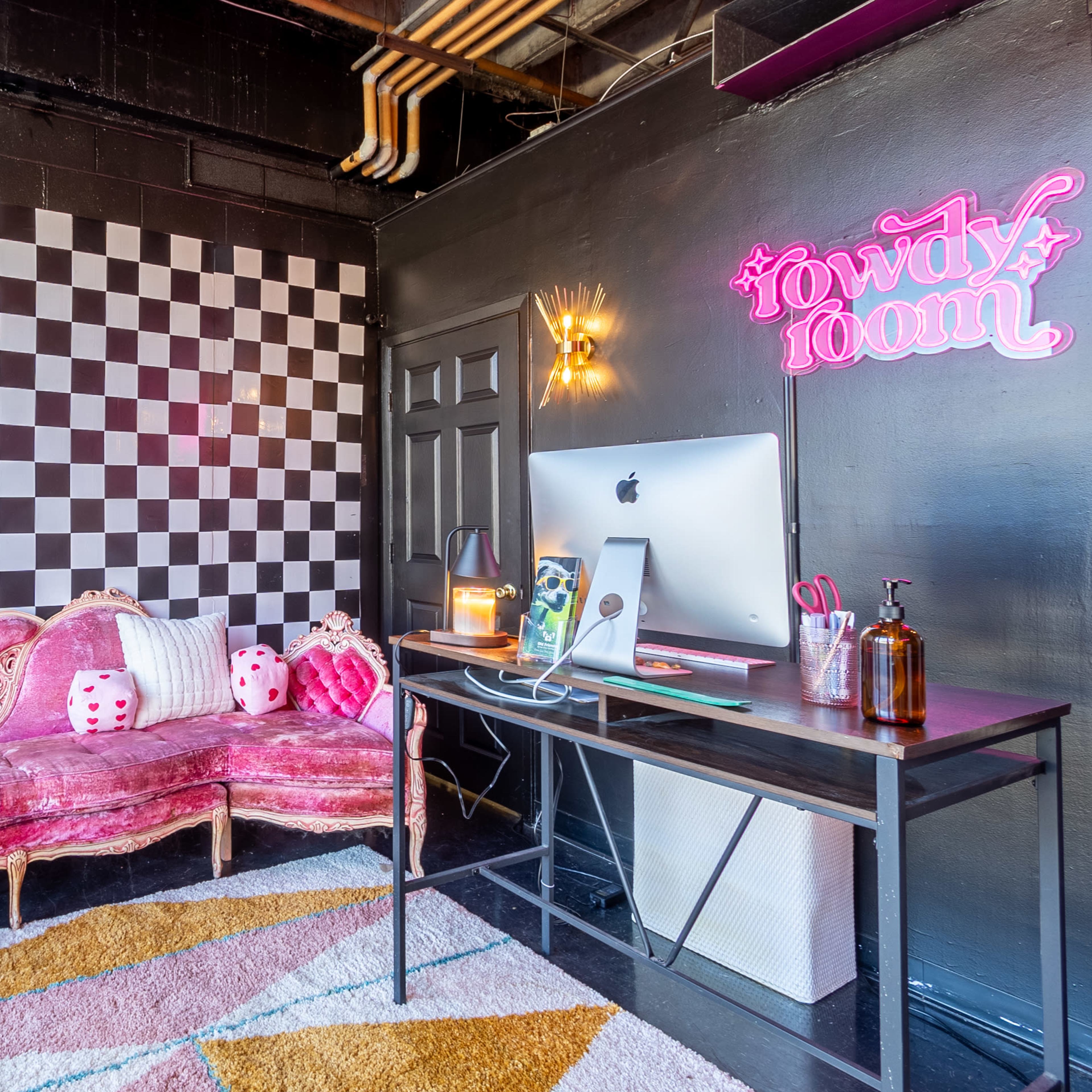A small room featuring a pink, tufted couch, a checkered wall, and a desk with a computer.