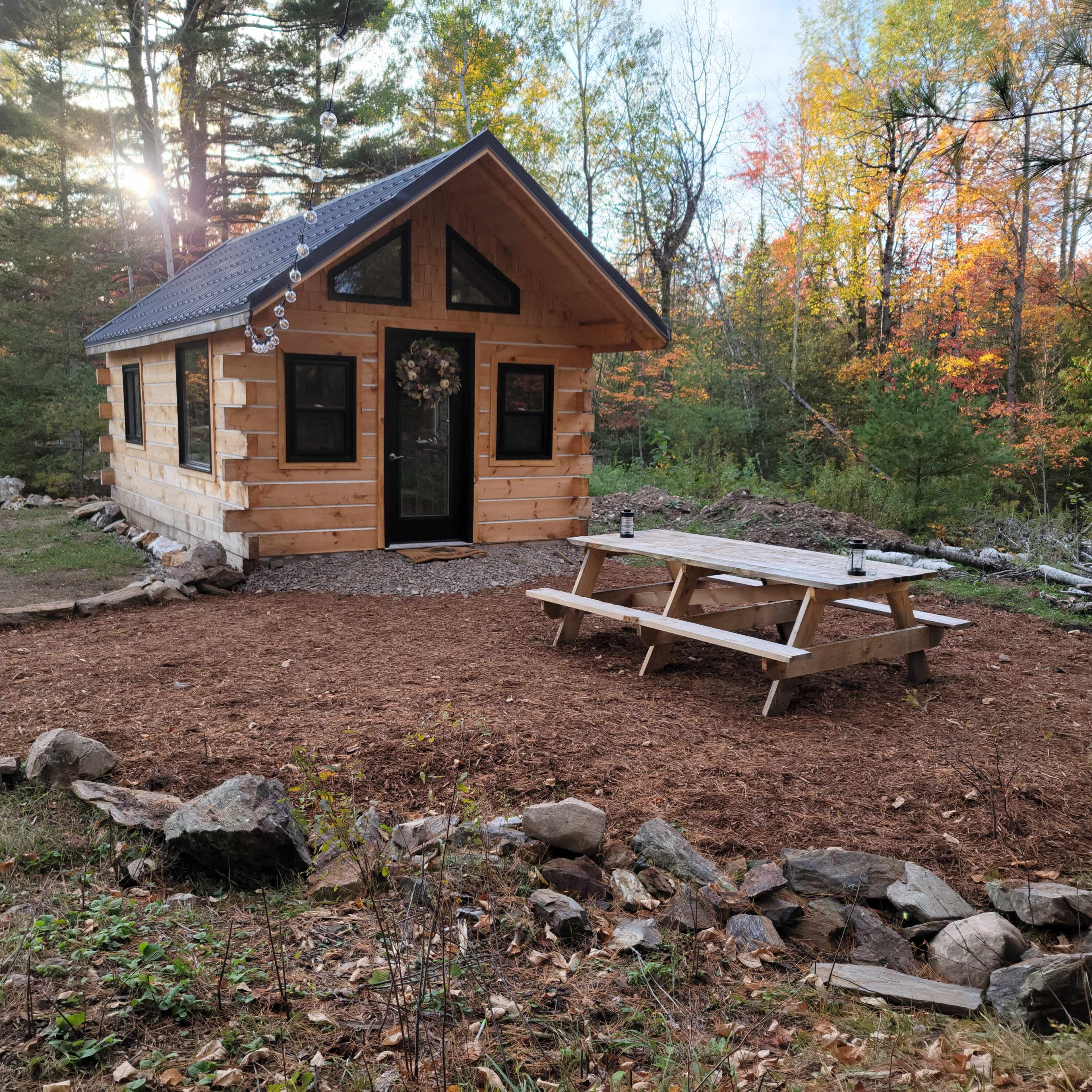 A small, log cabin with a front porch sits in a clearing surrounded by trees, with a picnic table placed nearby on a bed of mulch.