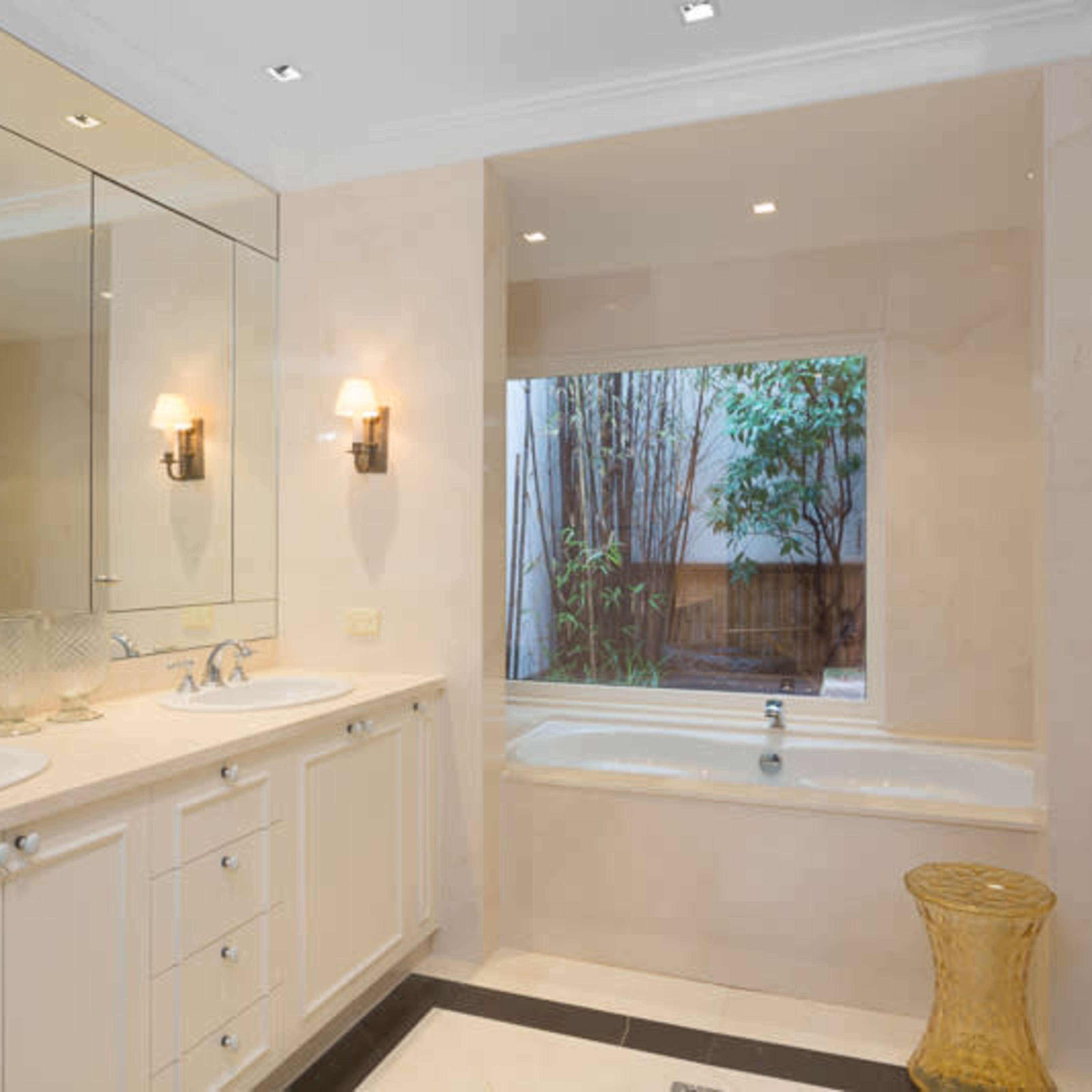 A modern bathroom with dual sinks, a freestanding bathtub, and large mirrors, illuminated by wall sconces and natural light from a window with greenery outside.