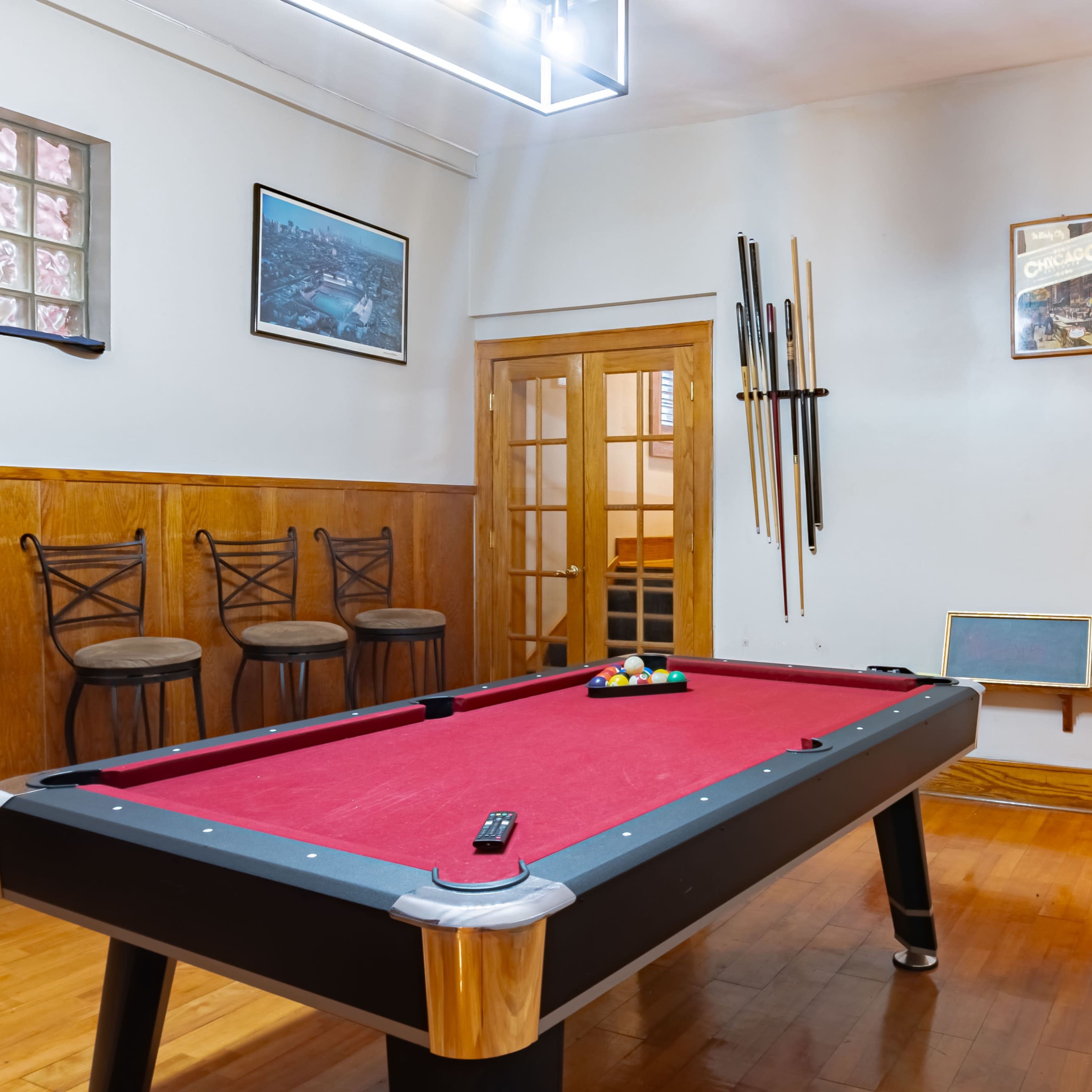 A billiards room featuring a red pool table, wooden chairs, and several cues mounted on the wall.